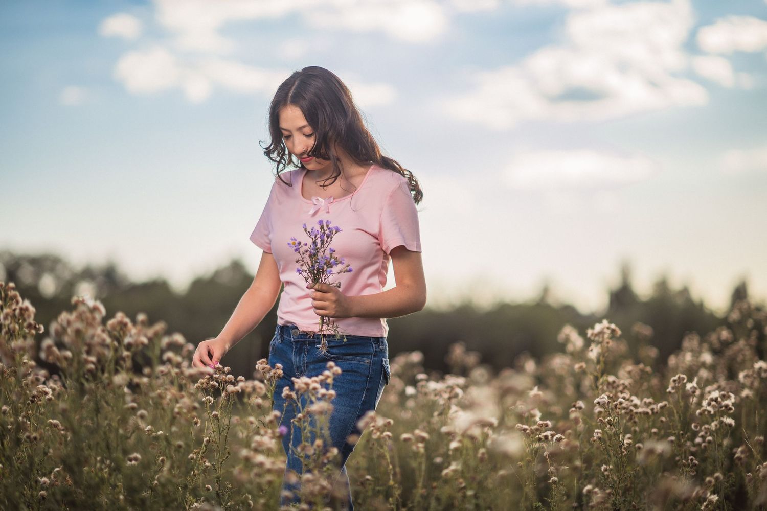 A person in a pink t-shirt strolls through a wildflower meadow at sunset holding delicate blooms.