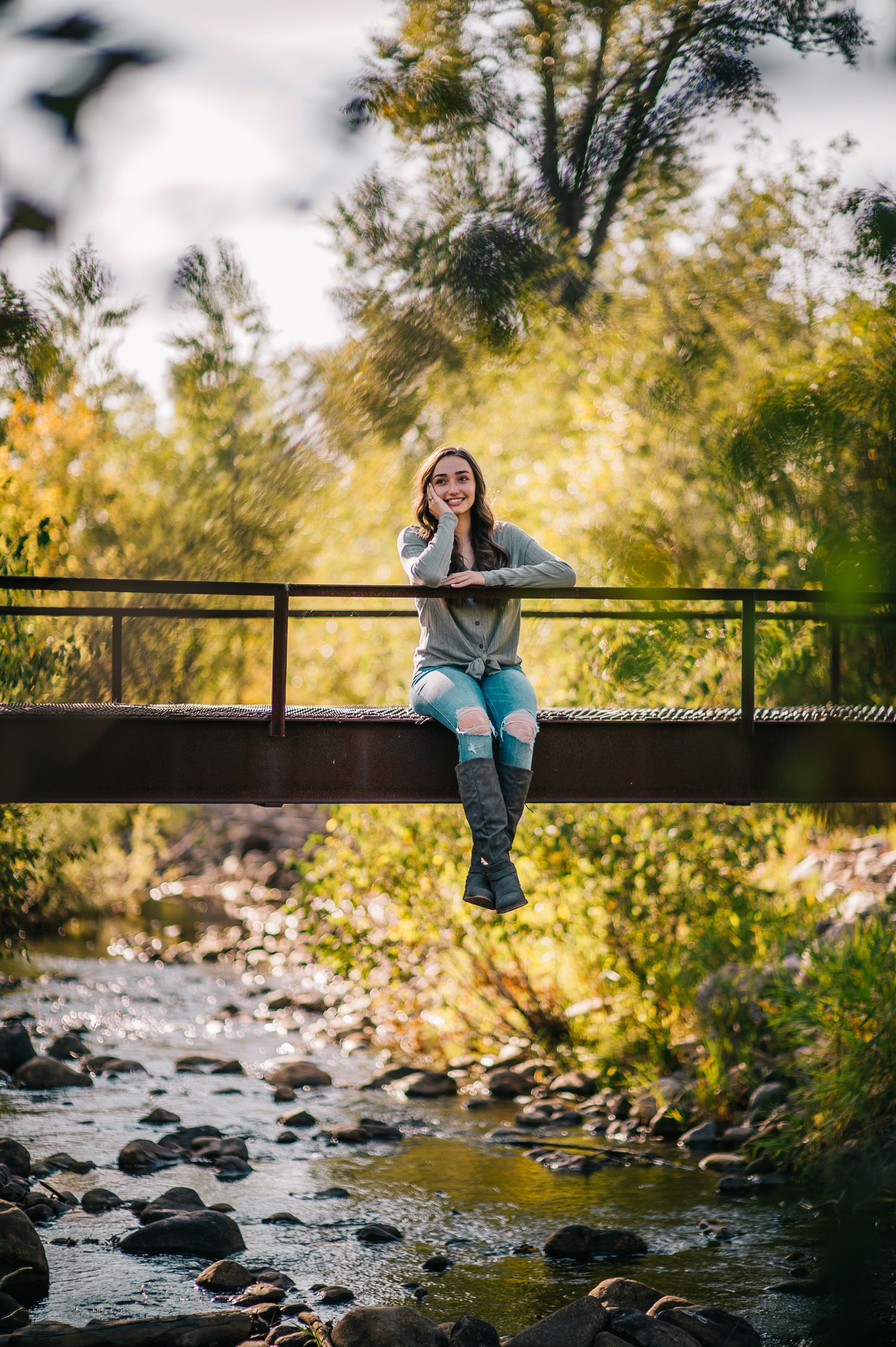 Person sitting on bridge railing over rocky stream during golden hour.