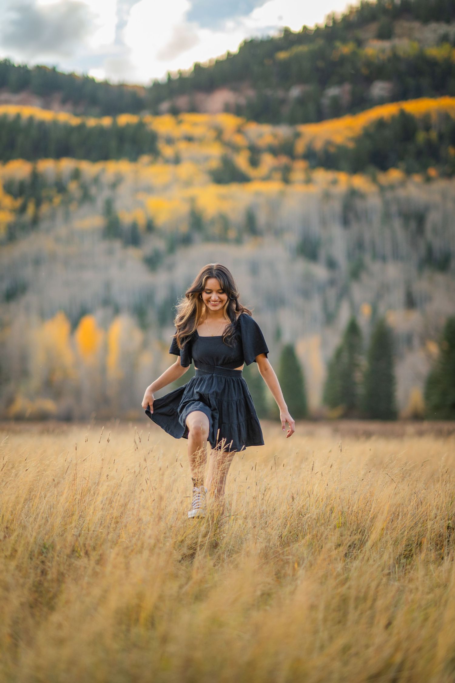 A person in a navy dress twirls through a golden mountain meadow with autumn aspens and evergreens in the background.
