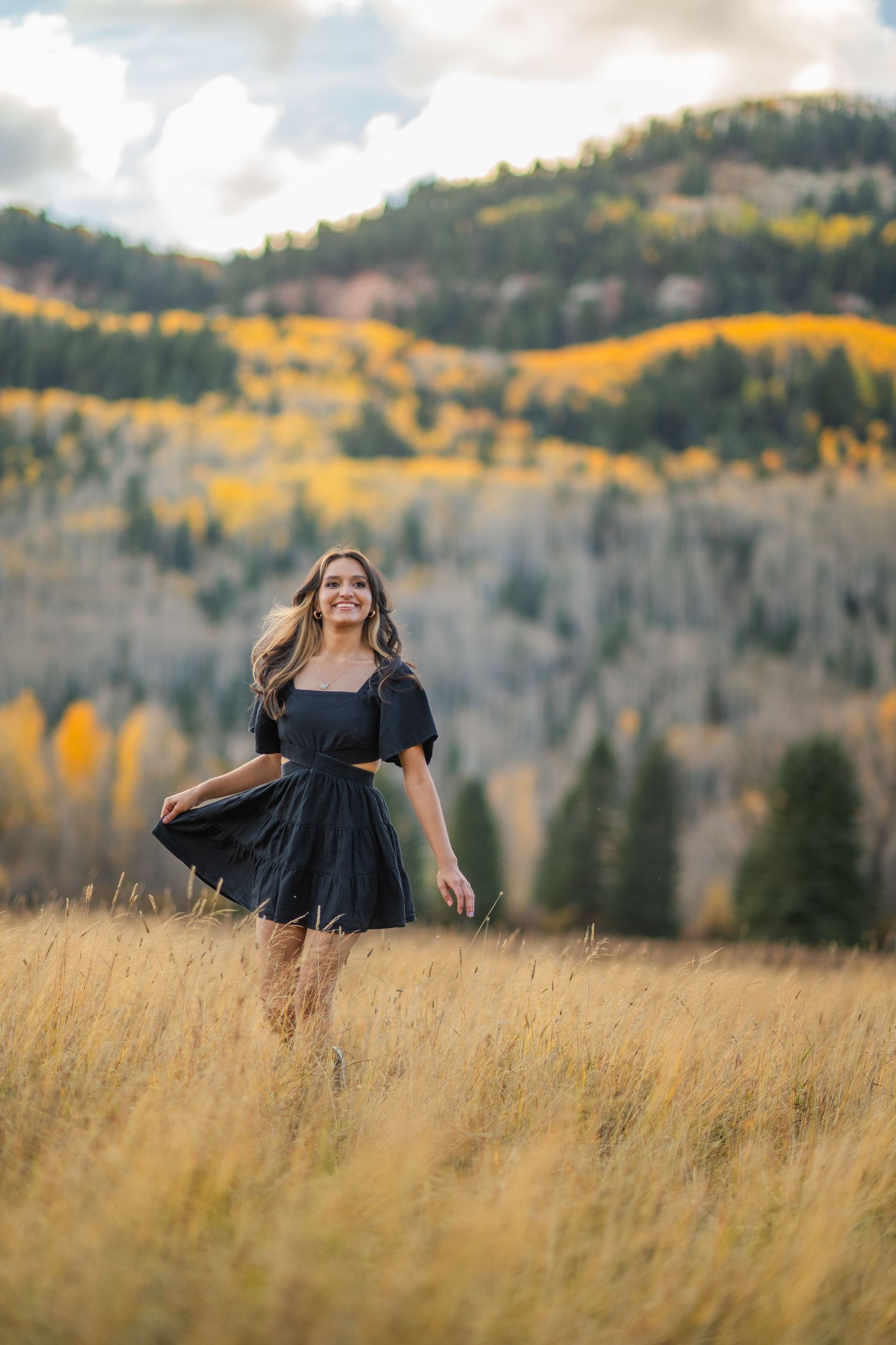 A person in a dark dress twirls joyfully in a golden mountain meadow with autumn aspens and evergreens in the background.