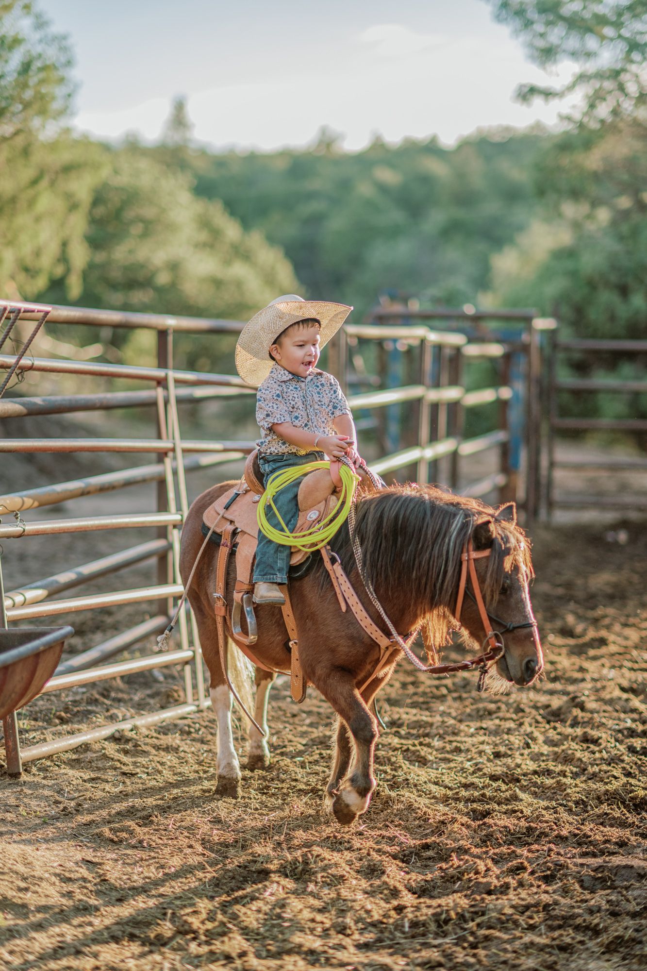Young child in cowboy hat riding a brown pony while holding a lasso in a ranch corral.