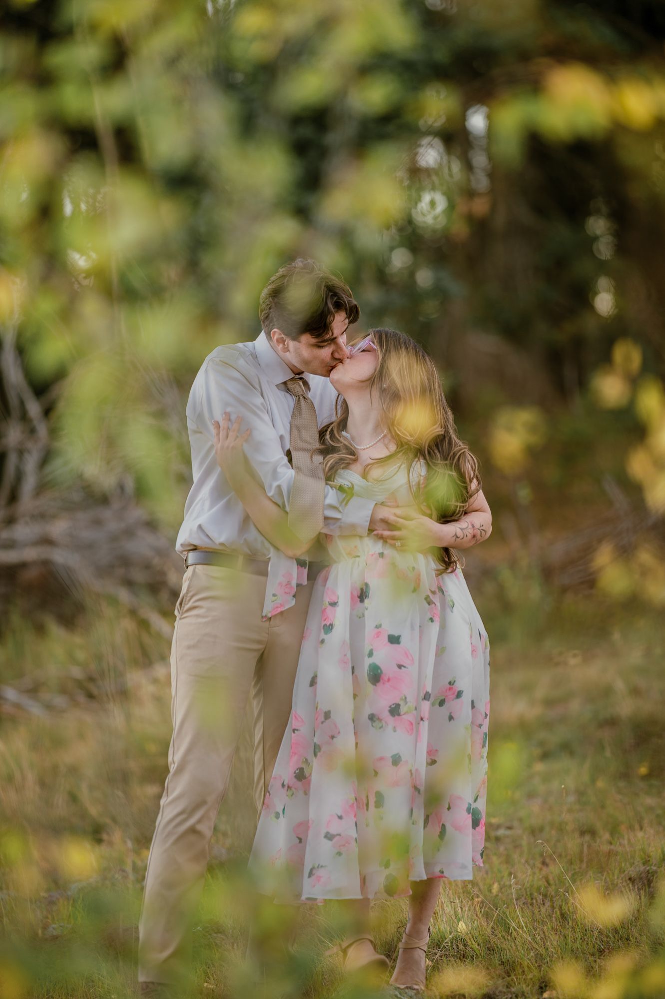 Couple kissing in a green forest during their engagement session near Chama, New Mexico, she wears a floral dress.