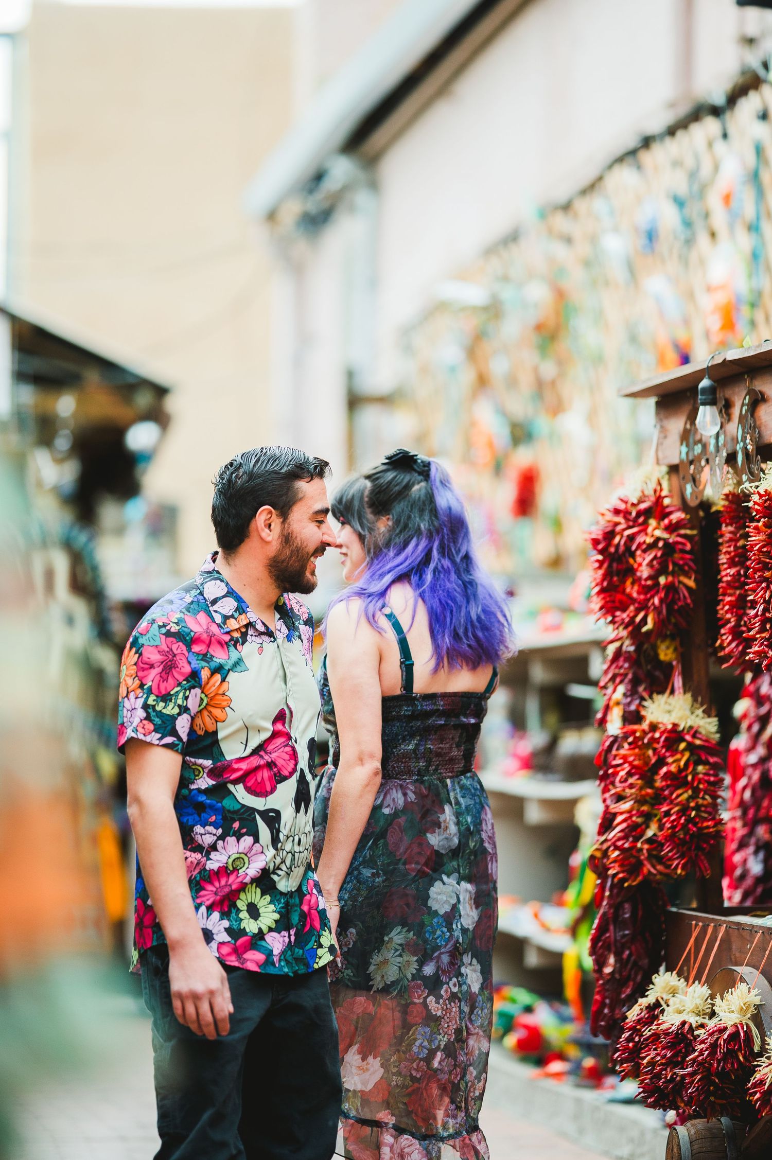 Couple in colorful floral outfits sharing a romantic moment at a vibrant outdoor market with hanging chili peppers.