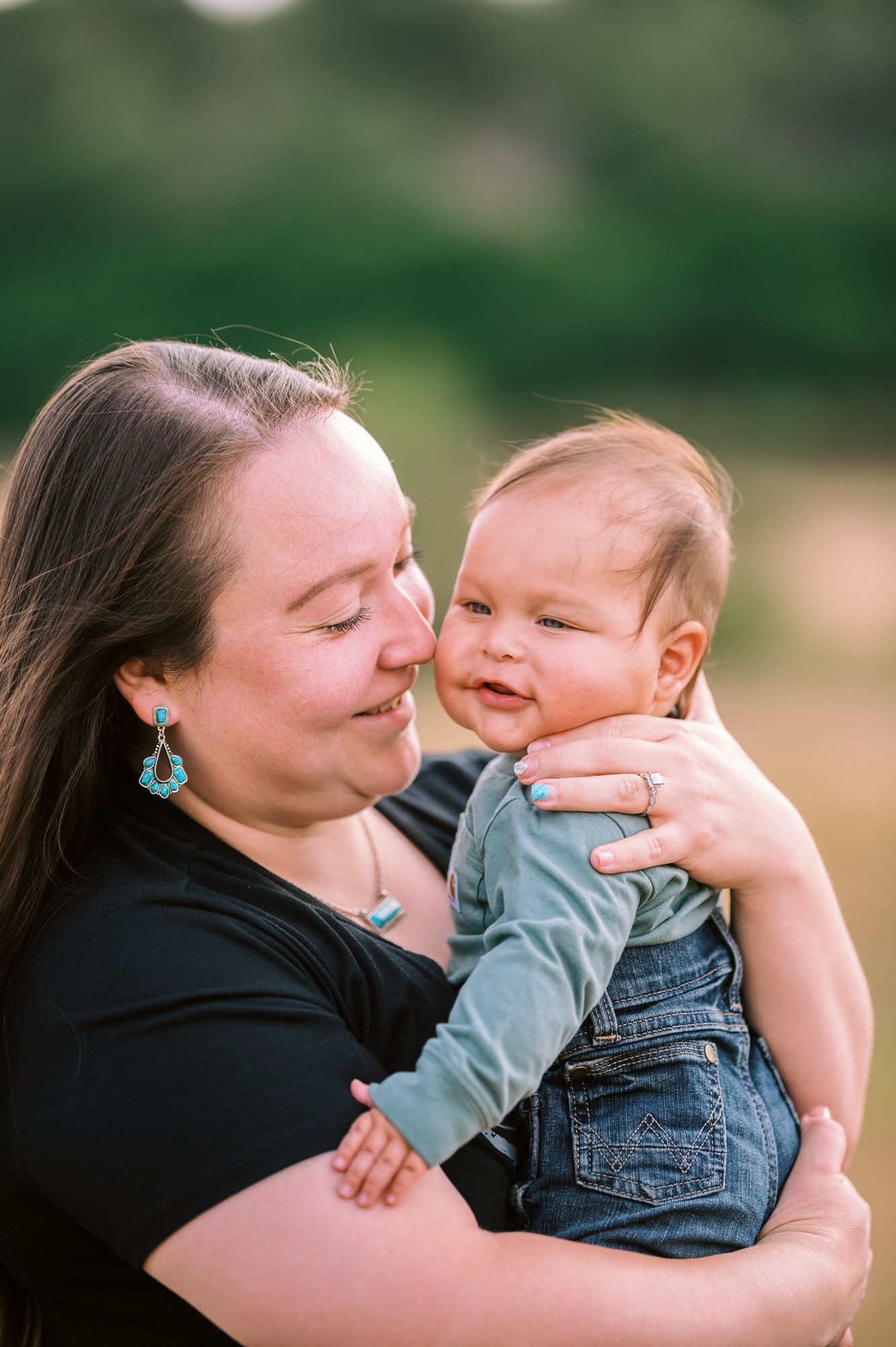 A mother tenderly holds her smiling baby dressed in denim overalls outdoors in a sunlit green field.