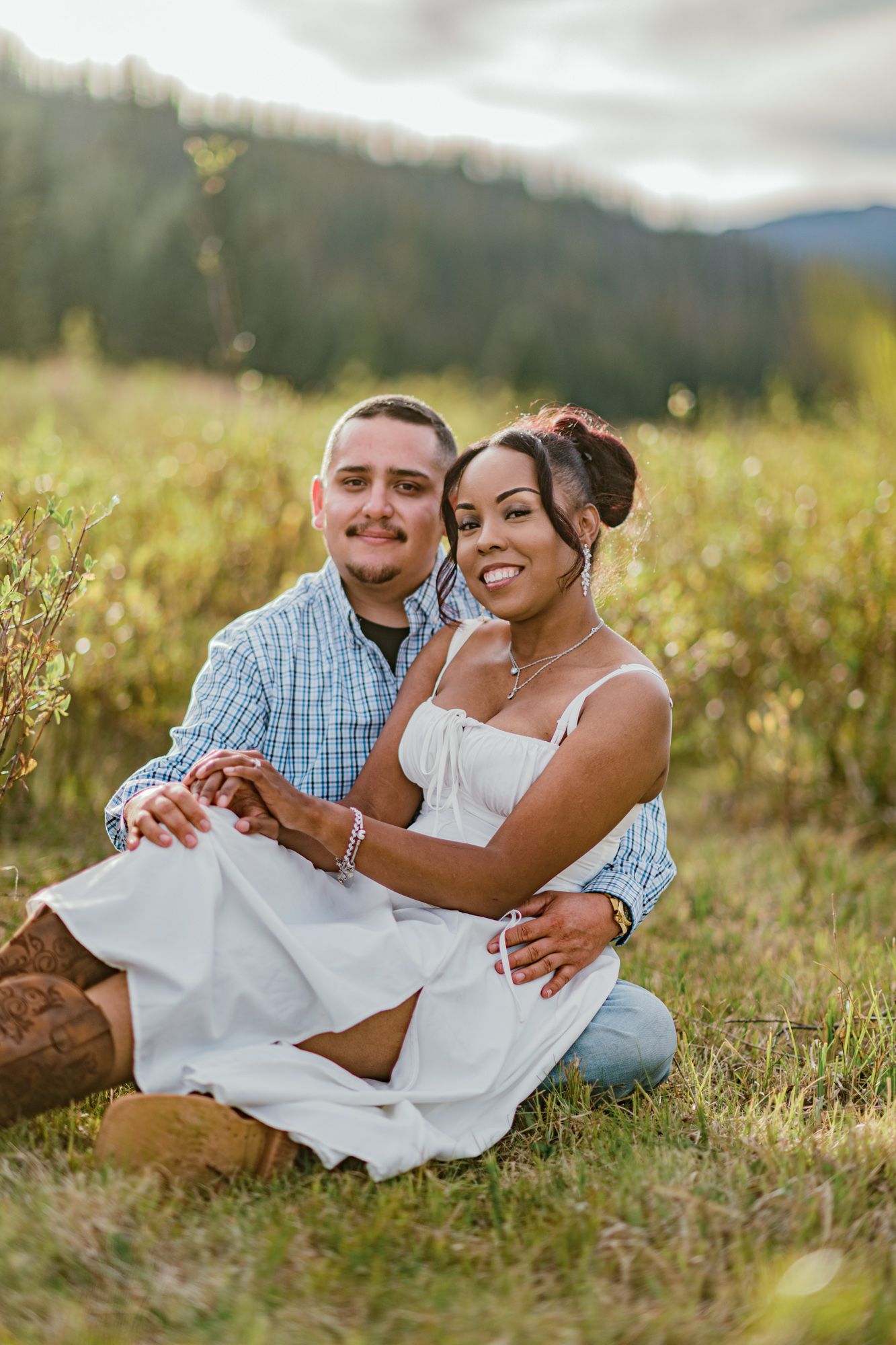 A smiling couple sits together in a sunny meadow, the woman in a white dress and the man in a plaid shirt.