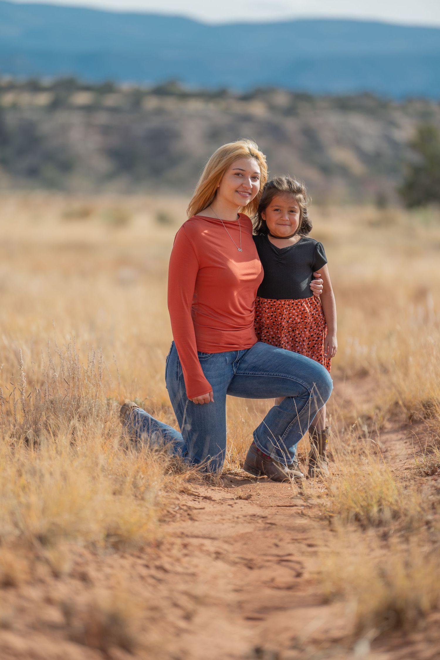 Two people wearing coral and navy clothing sit together in a desert landscape with mountains in the background.