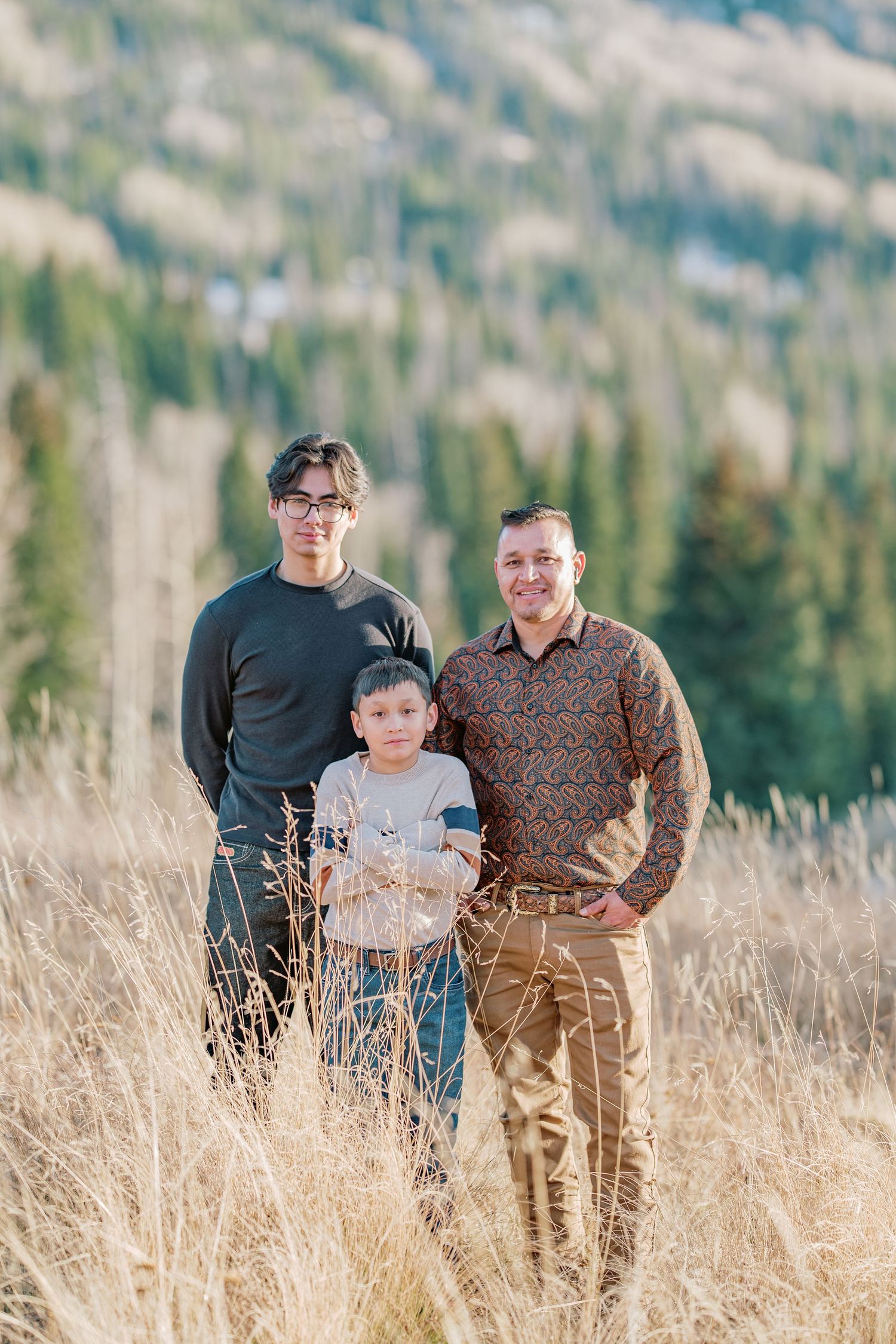A family of three stands together in tall golden grass with a mountainous forest landscape in the background on a sunny day.