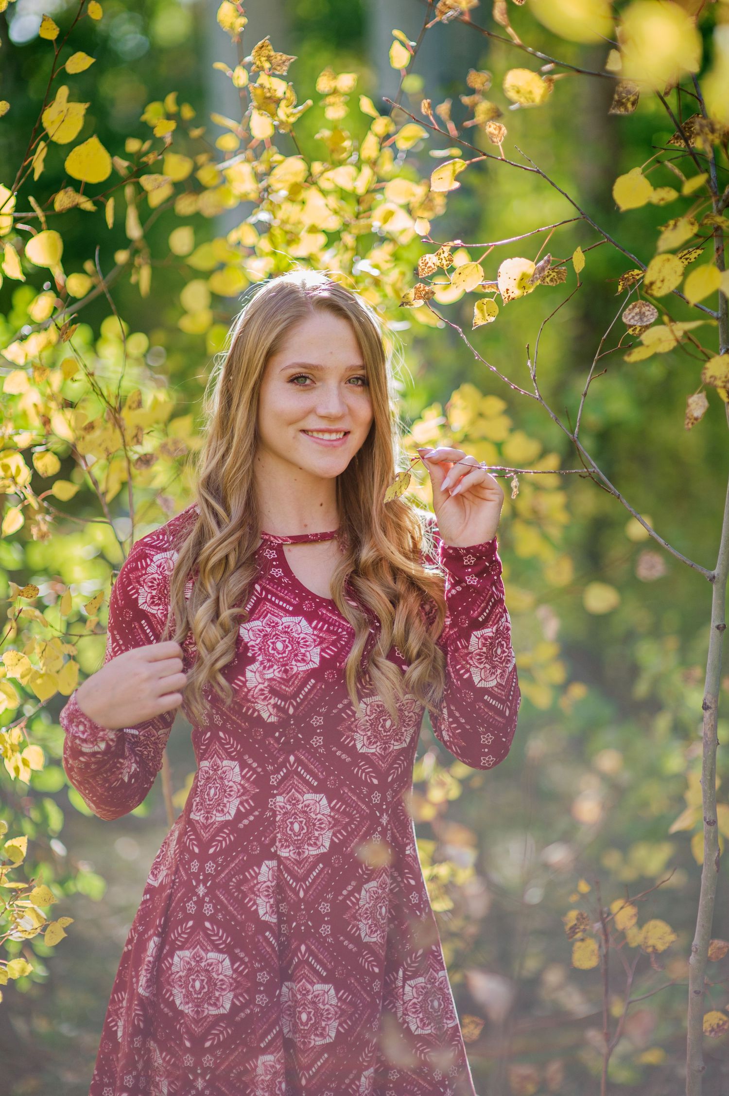 A woman in a burgundy patterned dress poses playfully against a backdrop of golden autumn foliage in dappled sunlight.