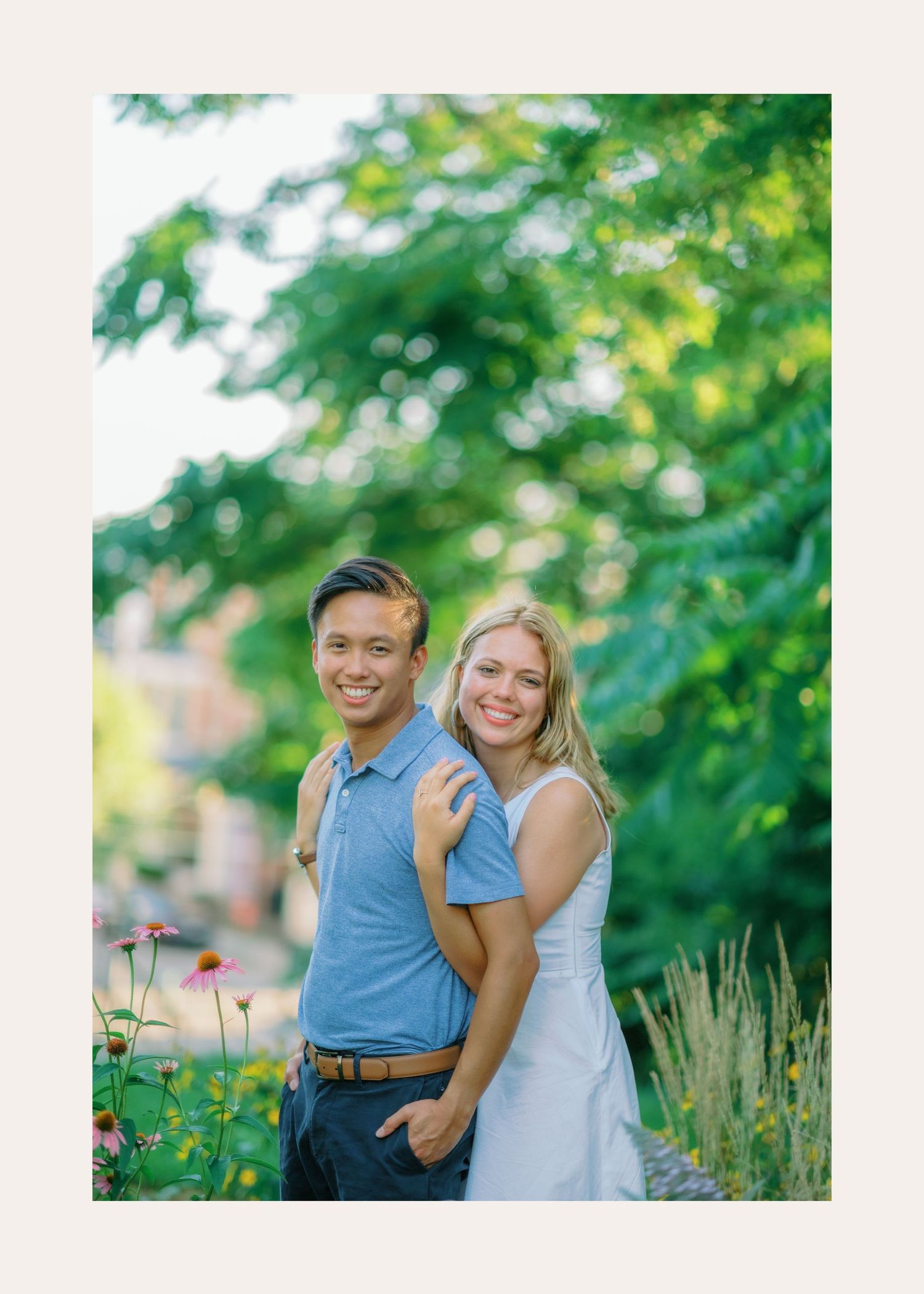 A couple poses together in a garden with blooming flowers and green trees in soft focus background.