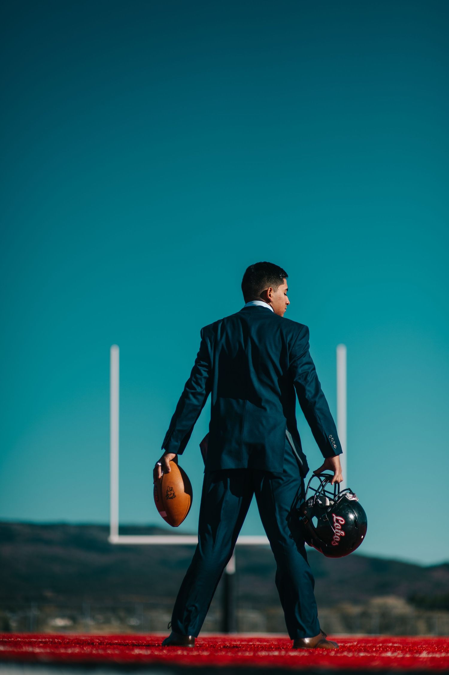 Man in suit holding football helmet and ball on field with goalposts against blue sky.