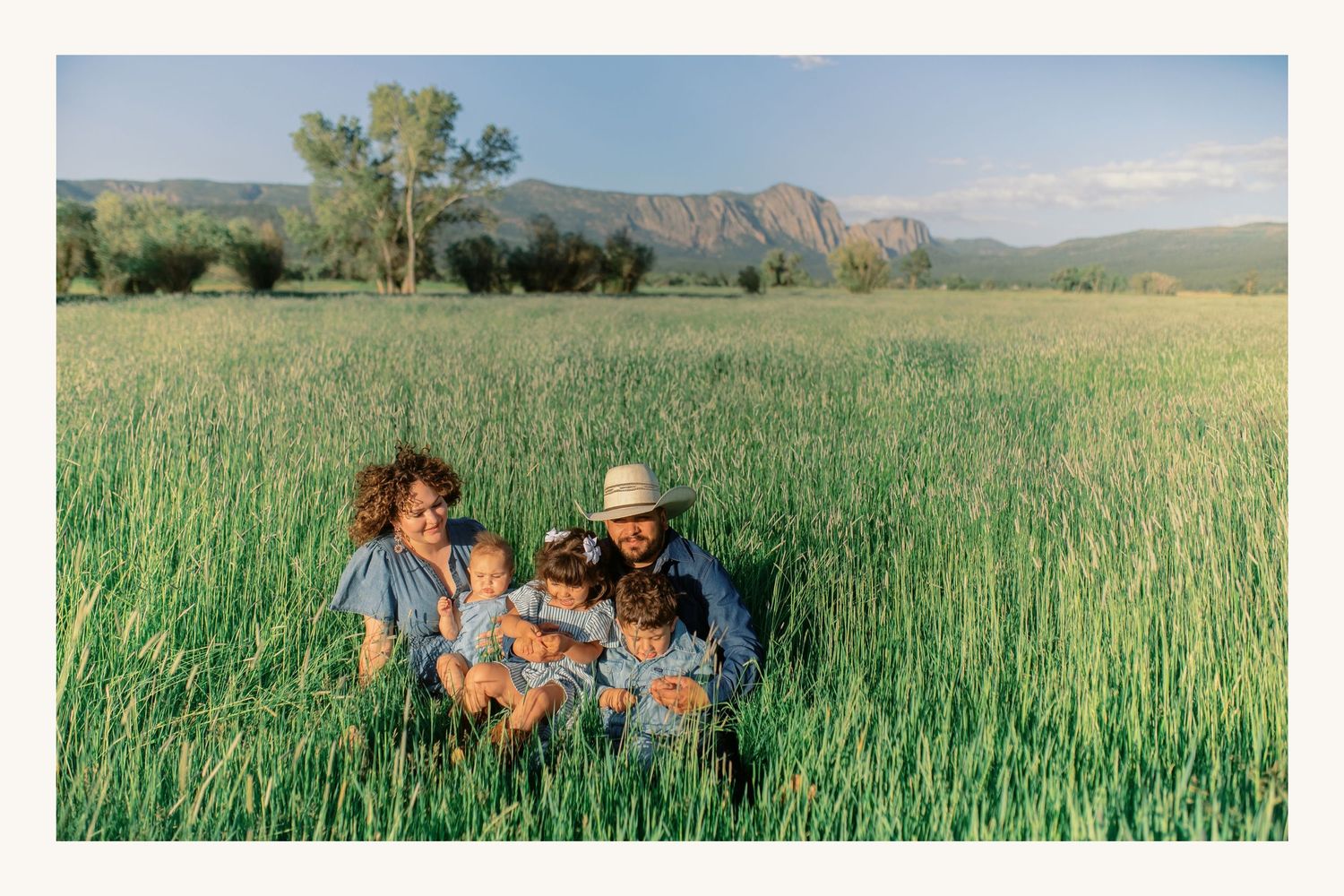 Family with baby sitting in lush green field with mountain backdrop on sunny day.