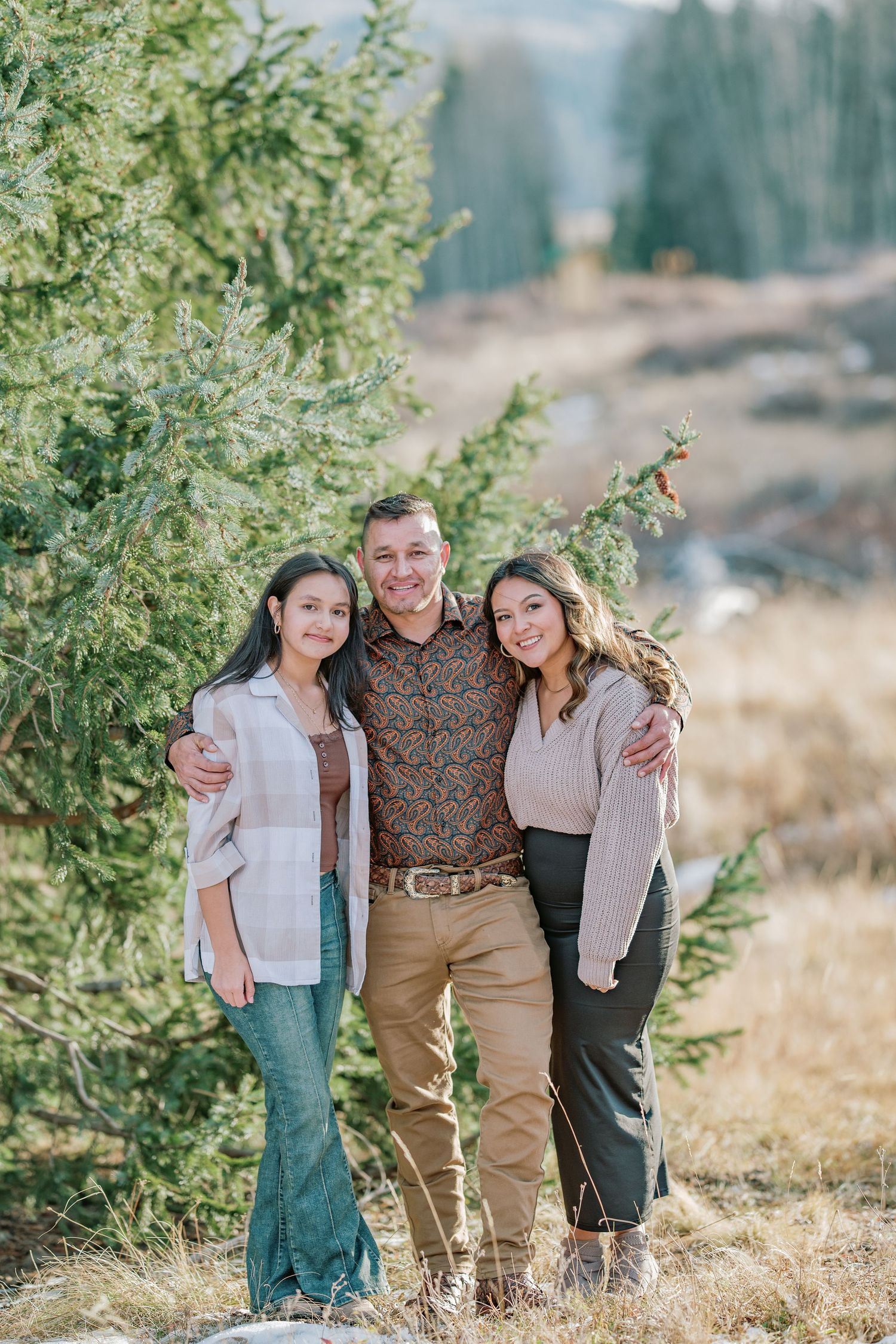 Three people stand together smiling in front of evergreen trees and mountain scenery on a sunny afternoon.