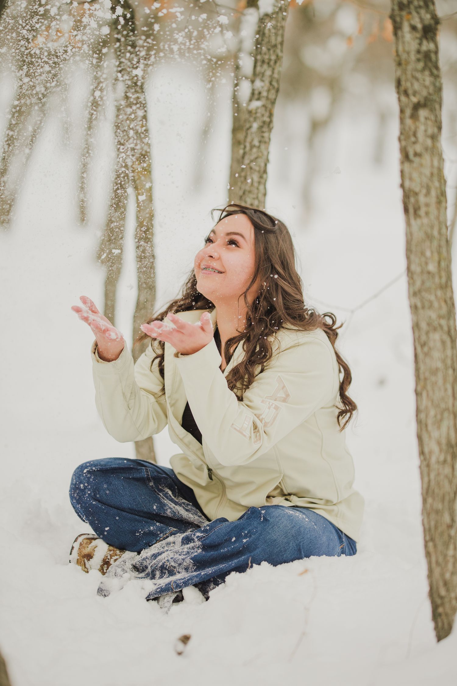 A person in a cream sweater and jeans sits cross-legged in snow while catching snowflakes in a winter forest.