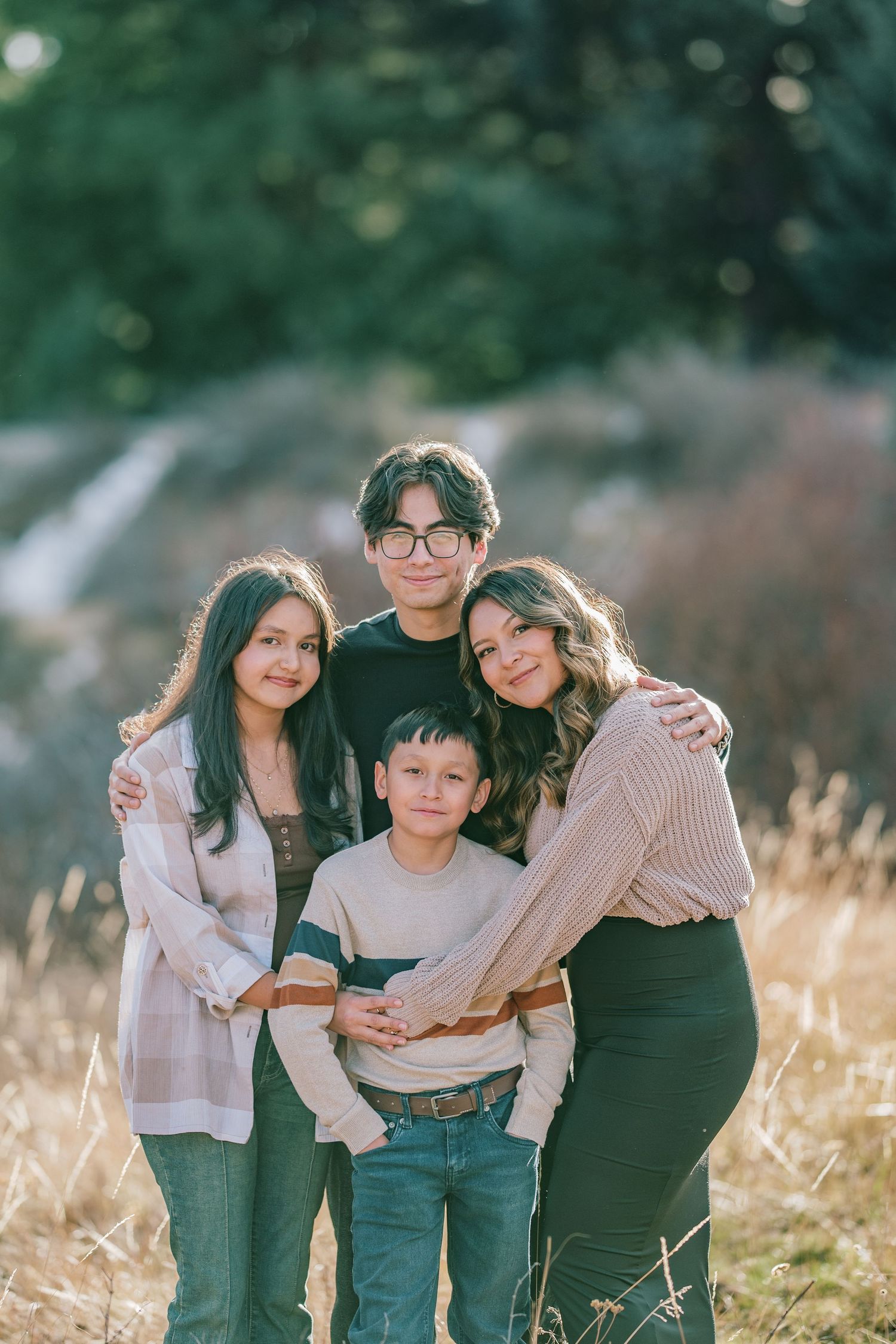 A family of four poses together in a sunny field with tall grass and green trees in the background.