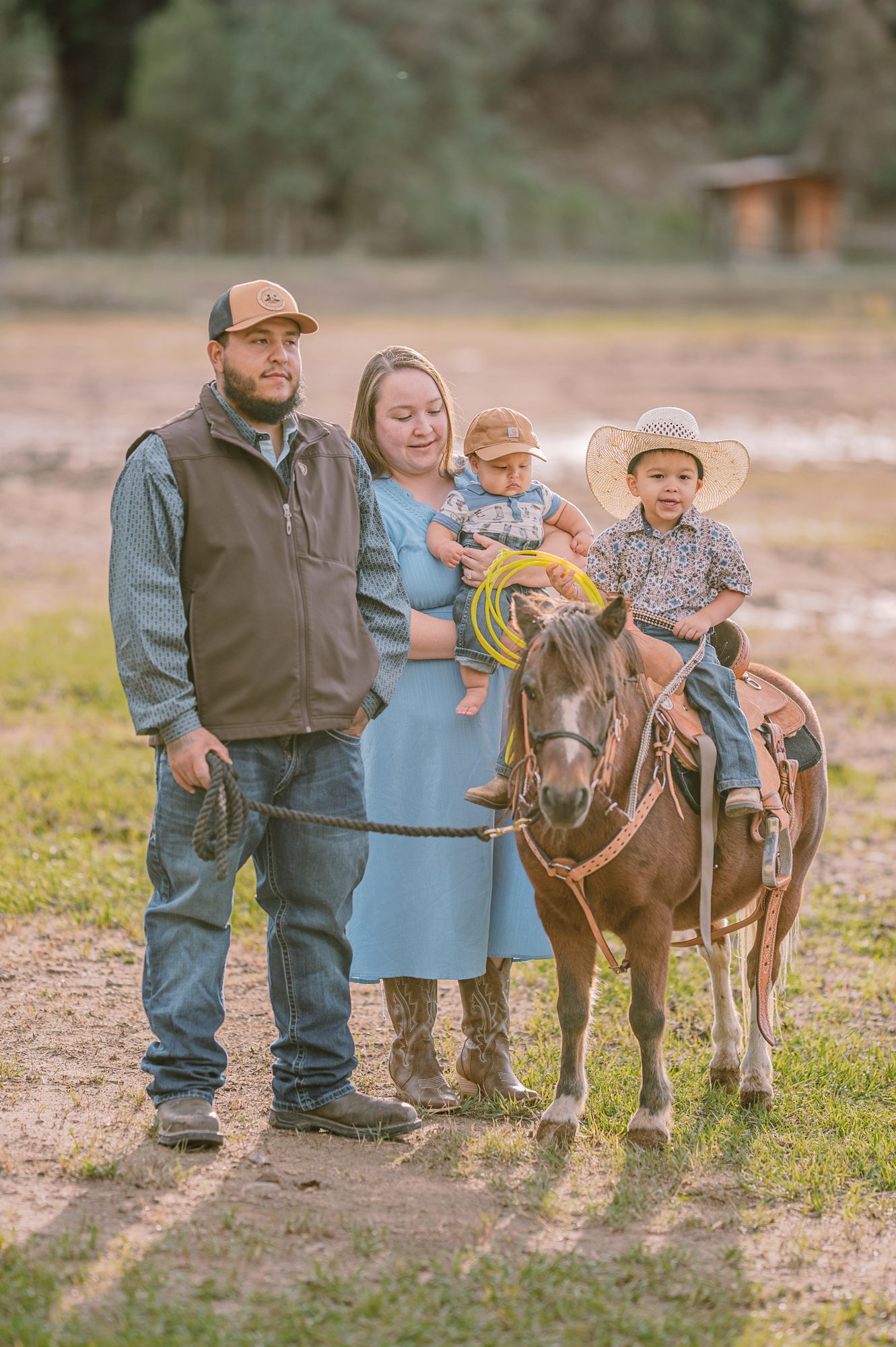 Family of four posing with a pony on a ranch, parents holding baby while young boy sits in saddle wearing cowboy hat.