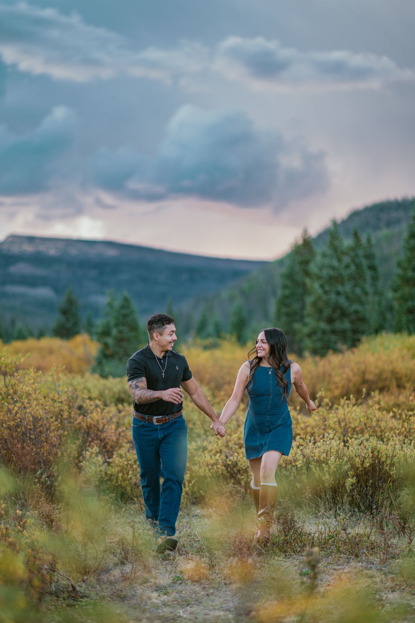 Couple holding hands walking through autumn mountain meadow with evergreen trees and dramatic cloudy sky at sunset.
