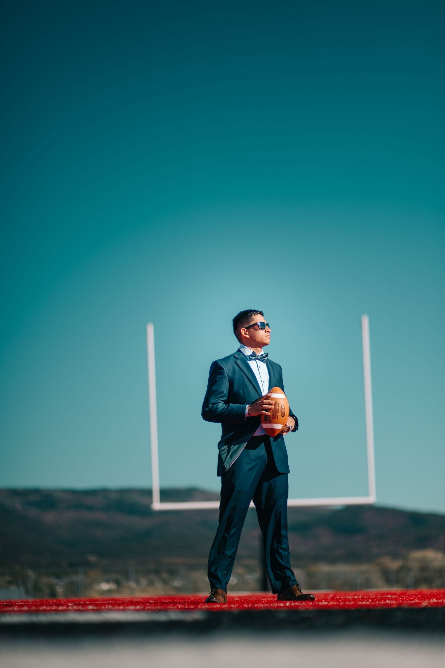 Young man in blue tuxedo holding football on field with goal post, wearing sunglasses under clear sky.