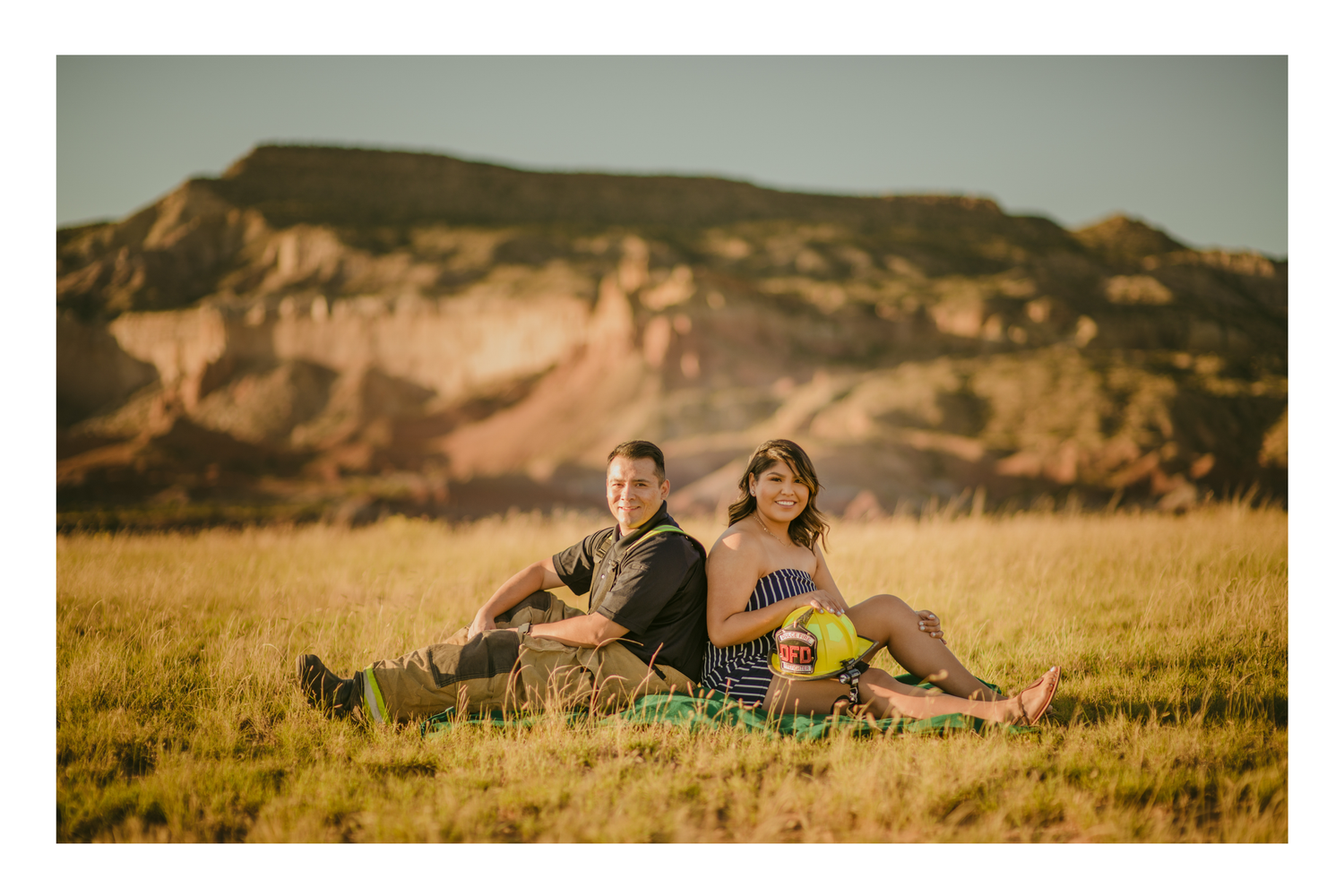 Couple sitting back-to-back in golden grass field with rocky badlands formation in background during sunset.