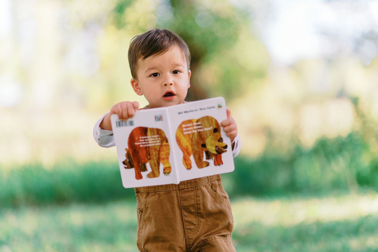 A child in brown overalls reads a picture book while sitting outdoors in a sunlit garden setting.
