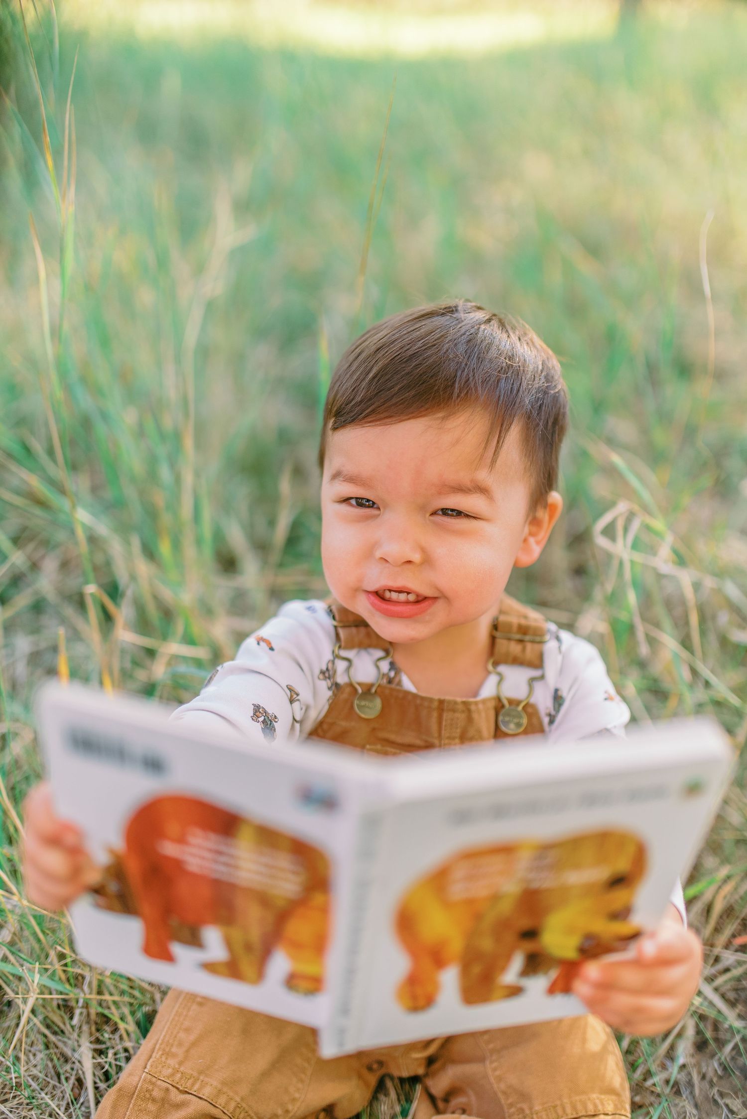A small child in brown overalls sits in grass while reading a picture book on a sunny day.
