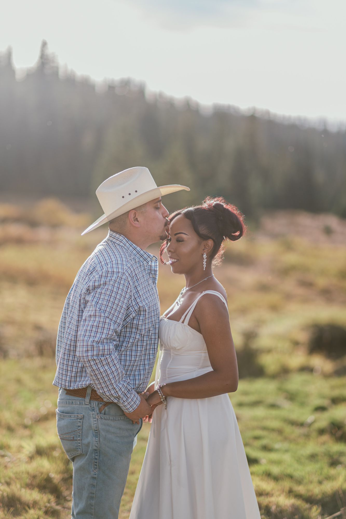 Interracial couple sharing a tender moment in a sunlit field, man in cowboy hat and plaid shirt, woman in white dress.