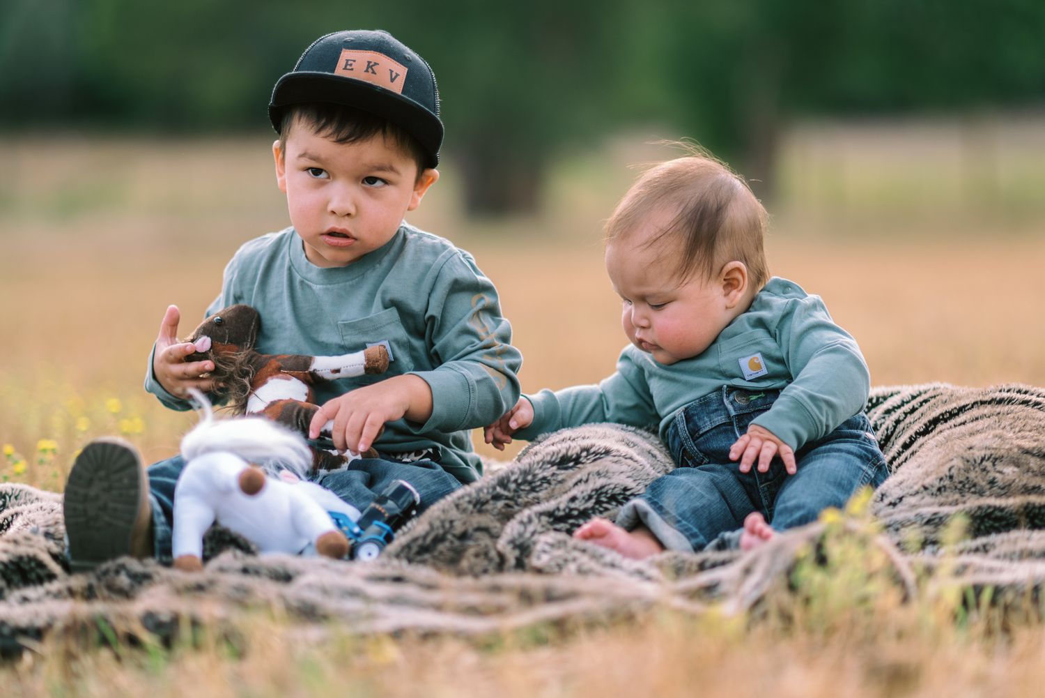 Two young children sitting outdoors on hay, playing with stuffed animal toys in a sunny field.