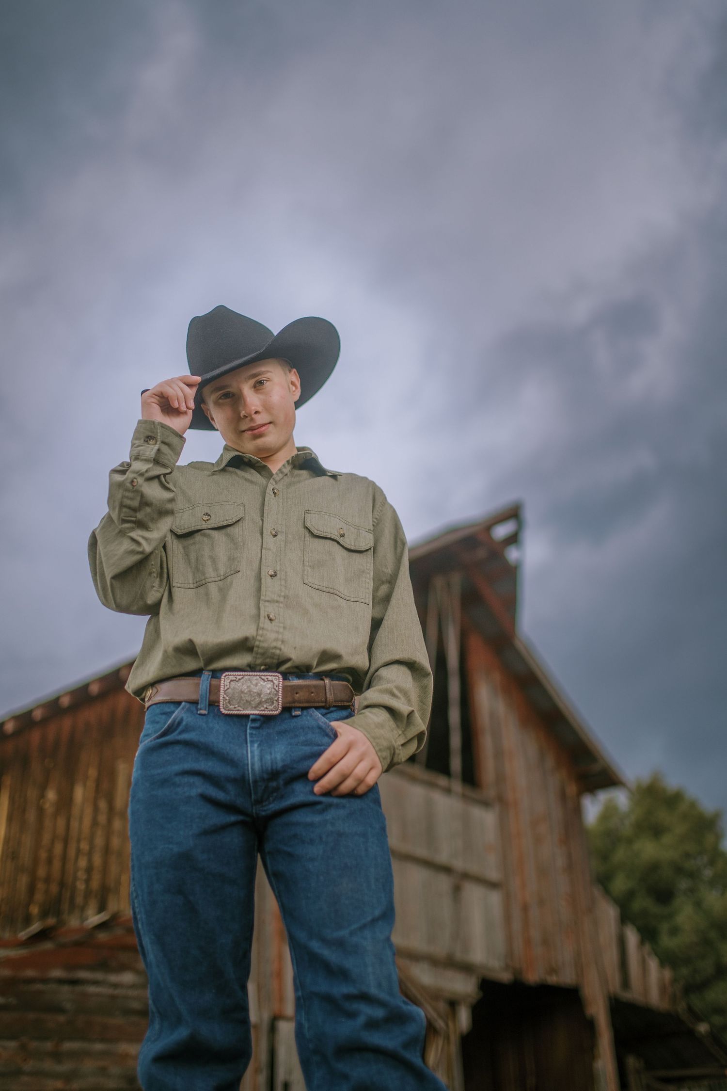 A person in western attire with jeans and cowboy hat stands in front of a rustic wooden barn under cloudy skies.