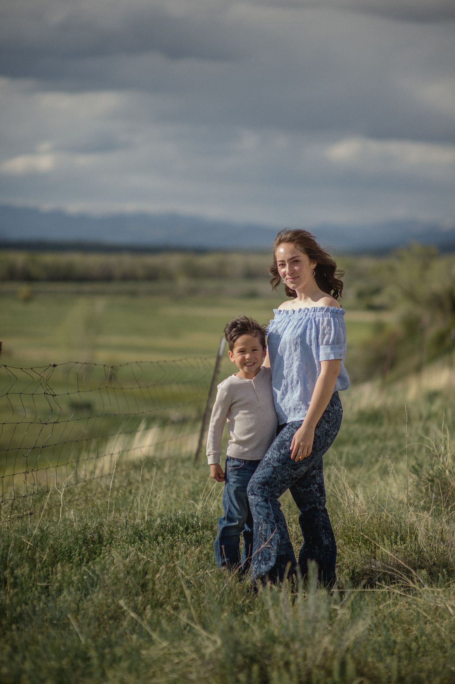 Two figures stand together in a grassy field with rolling hills and moody storm clouds in the background.