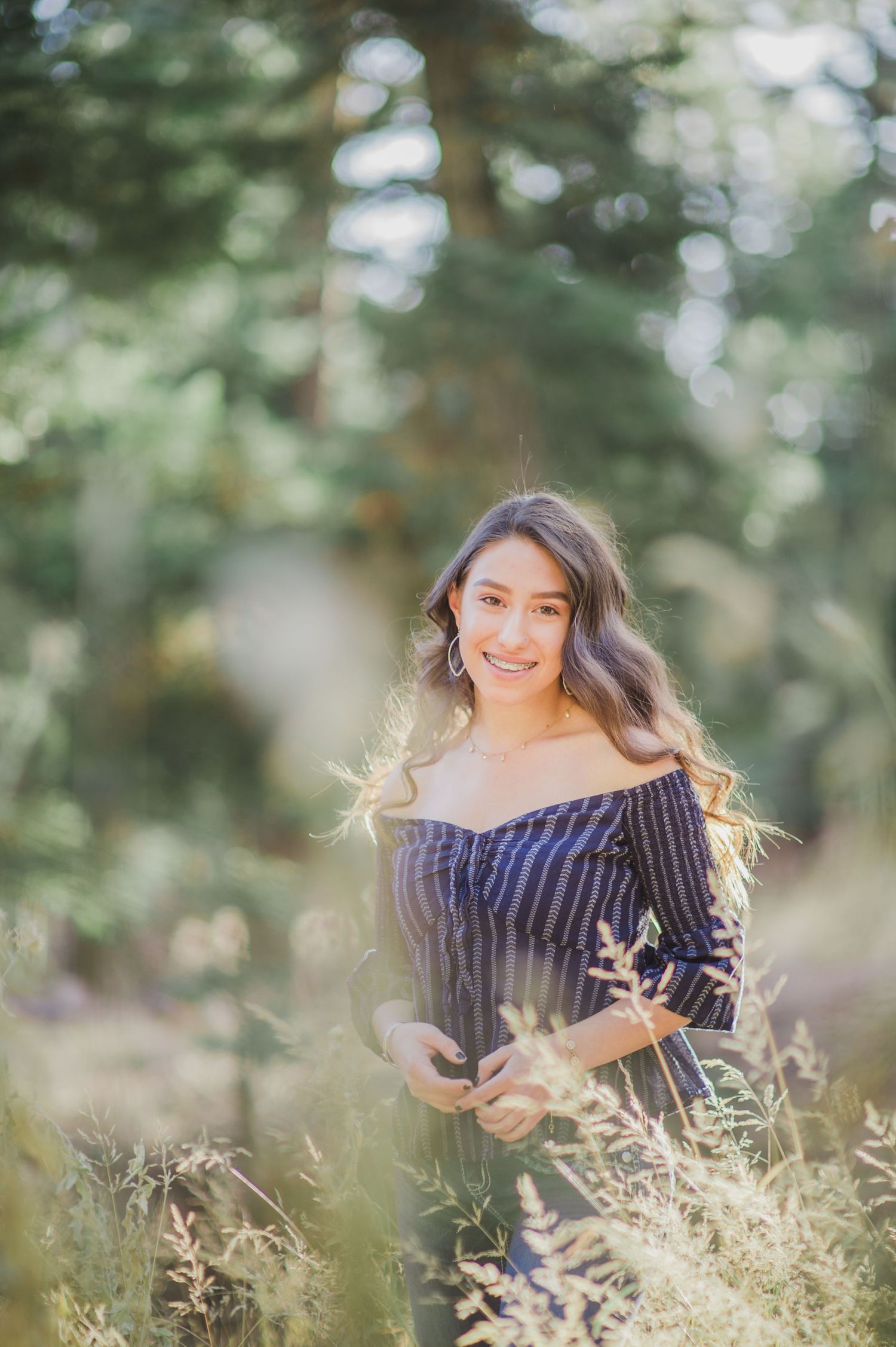 Young woman in striped off-shoulder top smiling in a sunlit forest meadow with tall grasses and green trees.