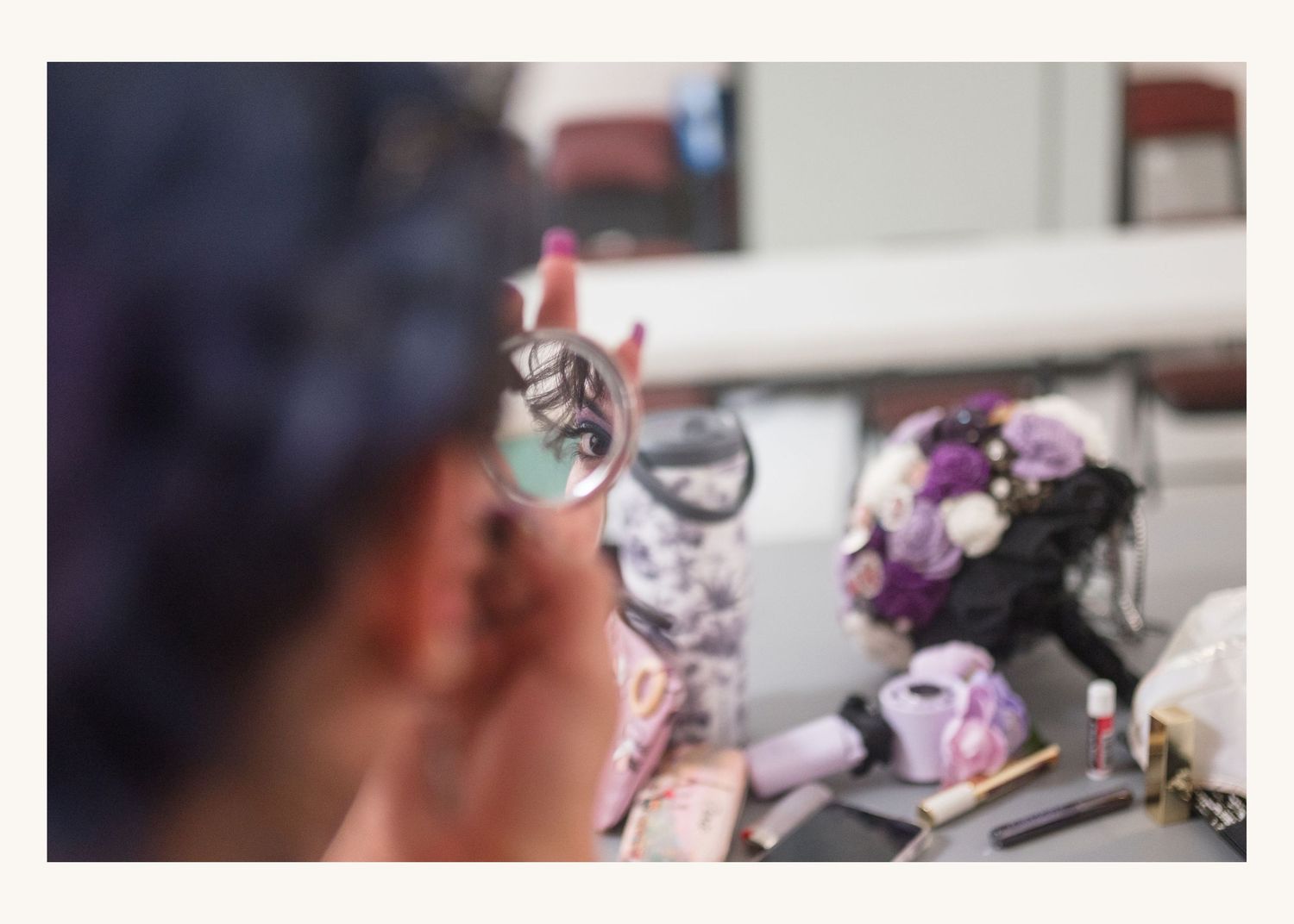 Bride checking makeup in small mirror at vanity with purple wedding bouquet in background.