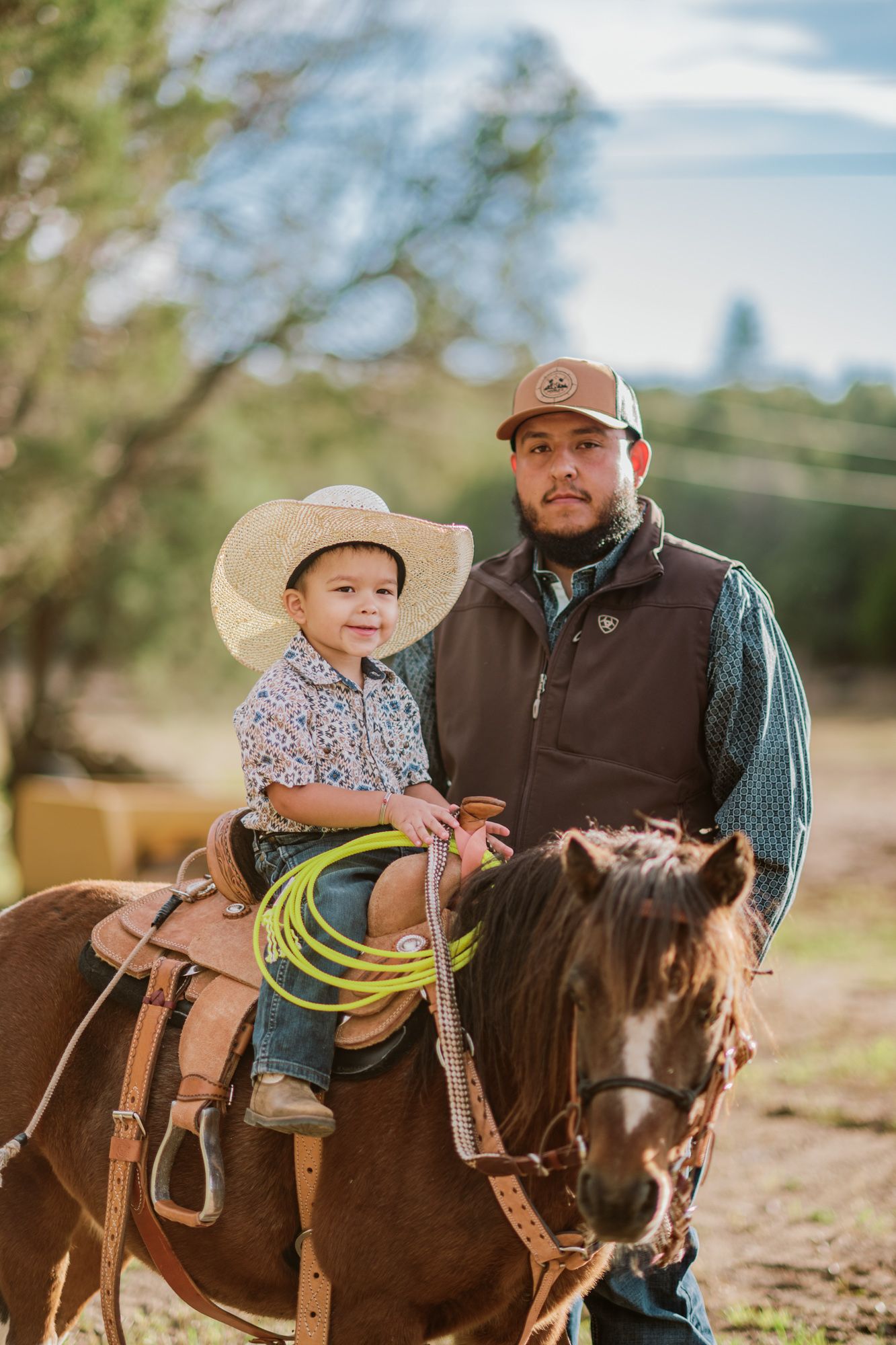 A man and young boy in cowboy hats sit together on a brown horse, with the child holding a lasso rope.