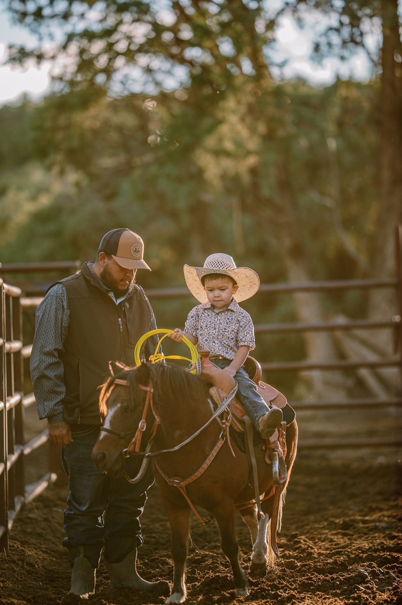 A young child in a cowboy hat rides a brown horse guided by an adult in a sunlit ranch corral.