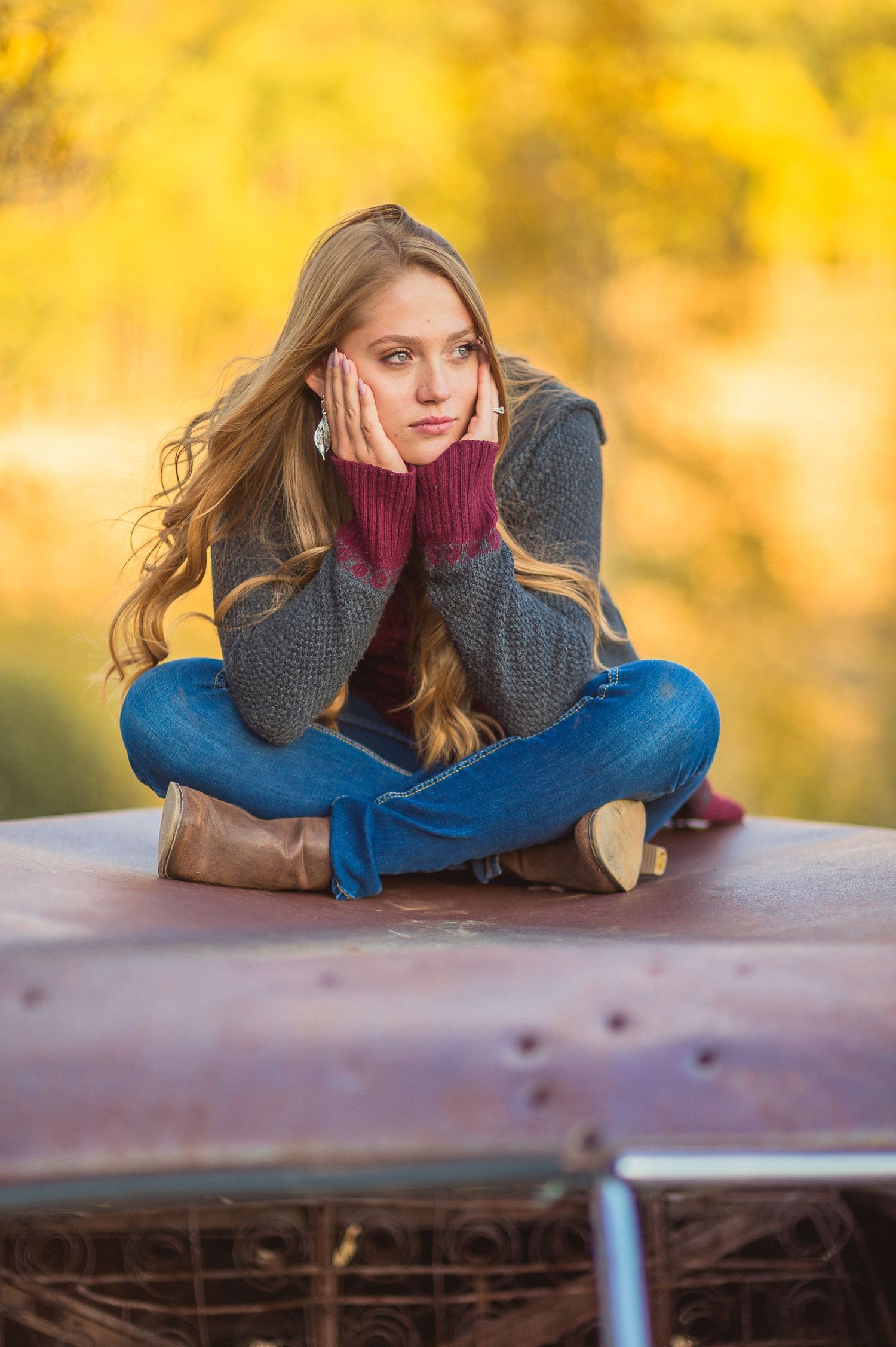 Portrait shot of someone seated on a car wearing cozy burgundy sweater and jeans against golden autumn background.