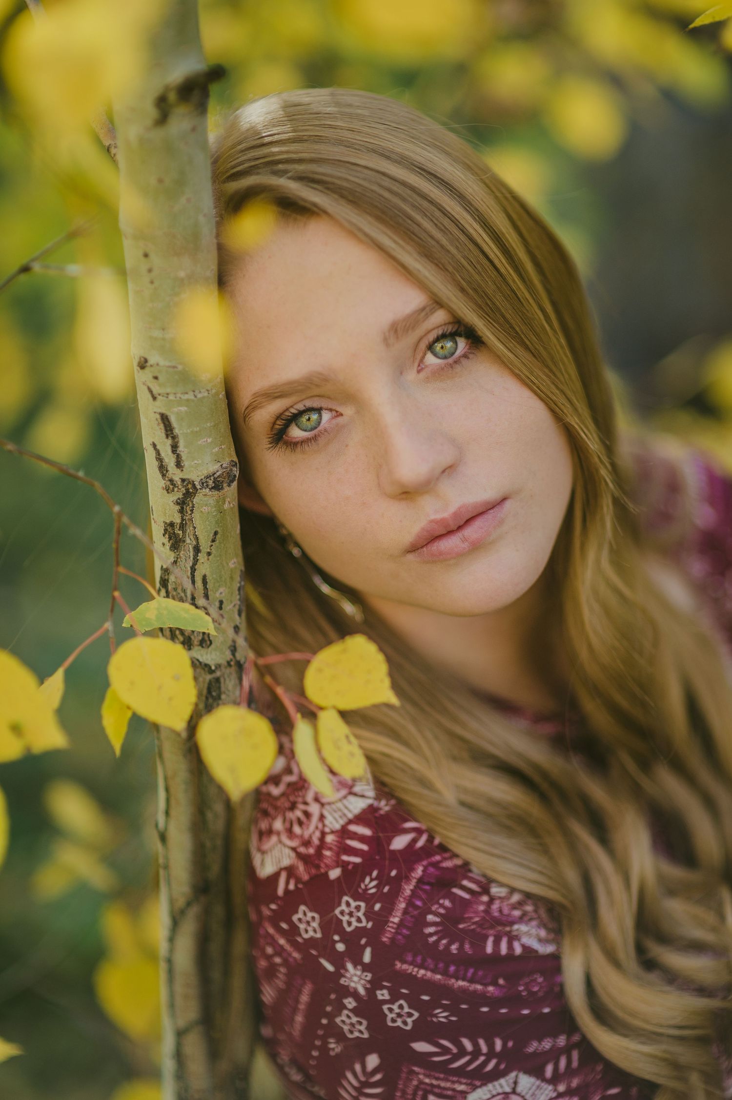 Portrait of someone with blonde wavy hair wearing a maroon top against autumn yellow leaves in nature.
