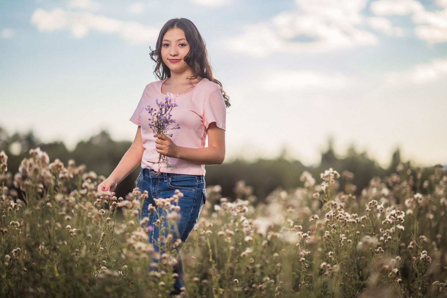 A woman stands in golden natural light among white wildflowers in a serene meadow at sunset.