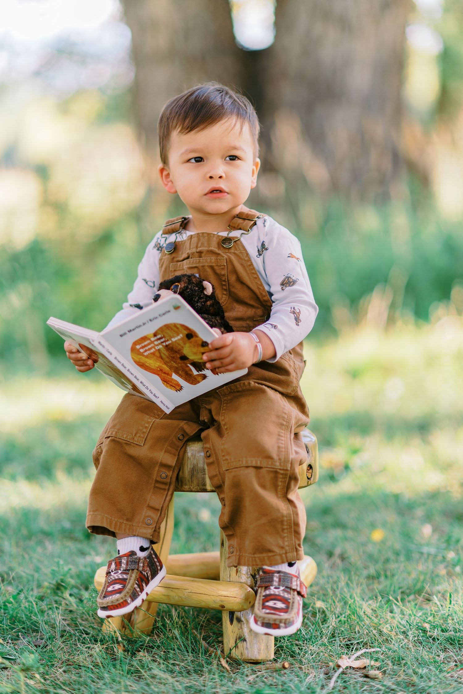 A toddler wearing brown overalls sits in grass while holding a book outdoors in a natural setting.