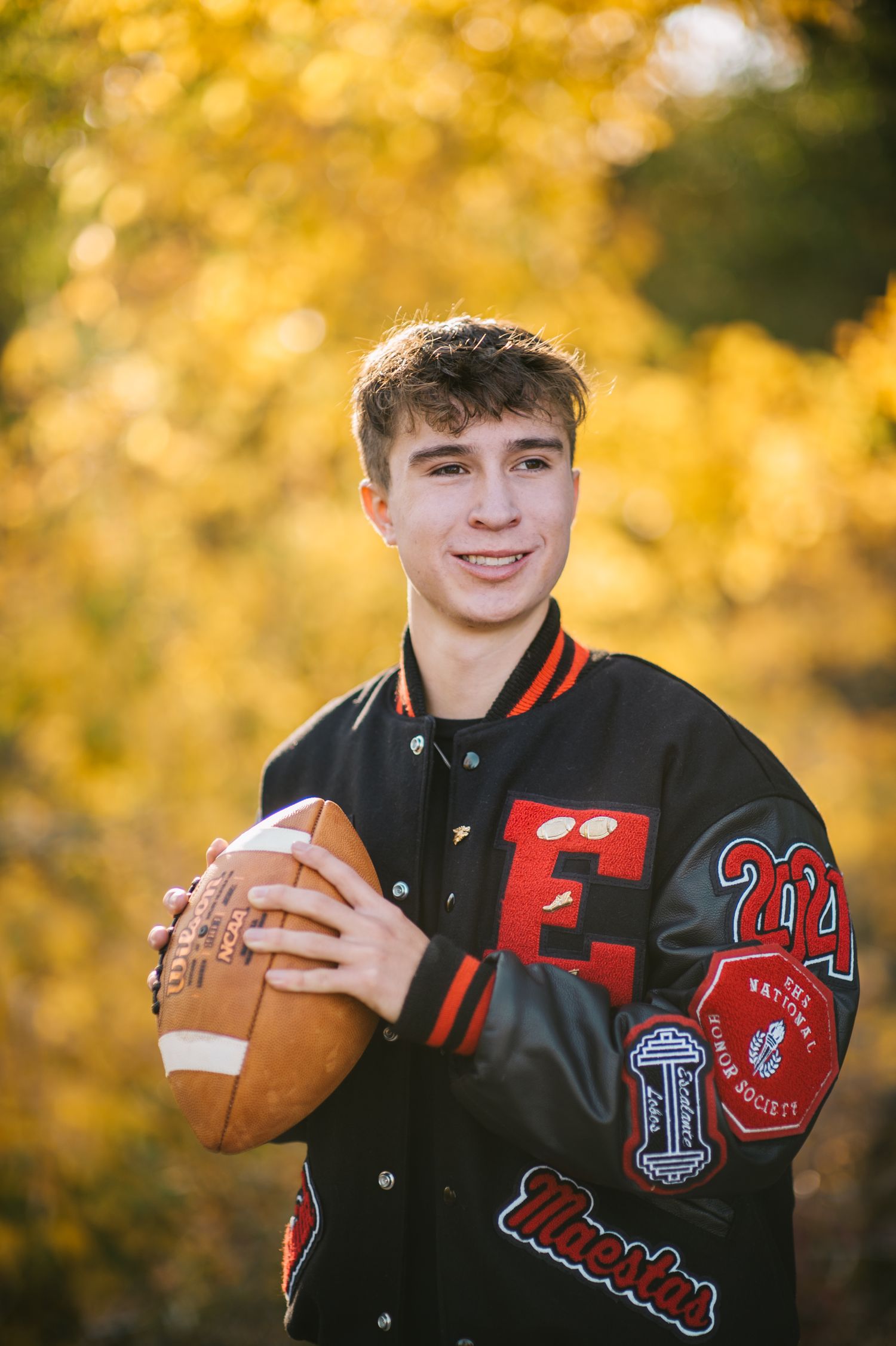 Athletic letterman jacket and football shown in autumn portrait session with golden background.