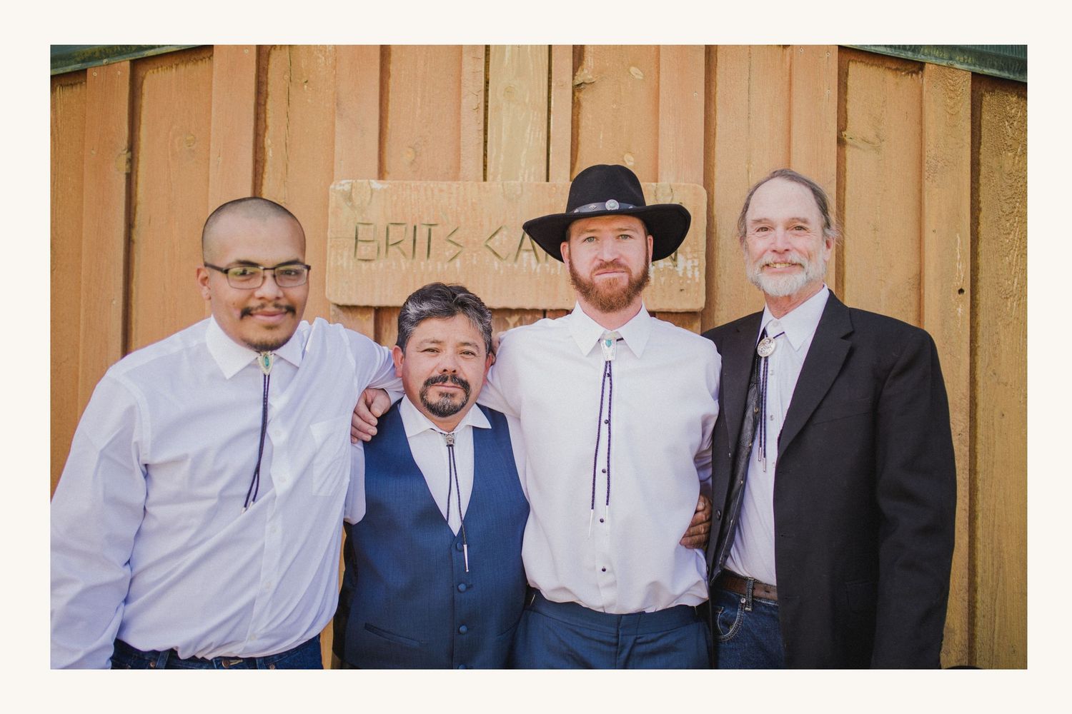 Four men smiling together outdoors near a wooden wall with a sign, dressed in formal and semi-formal attire.