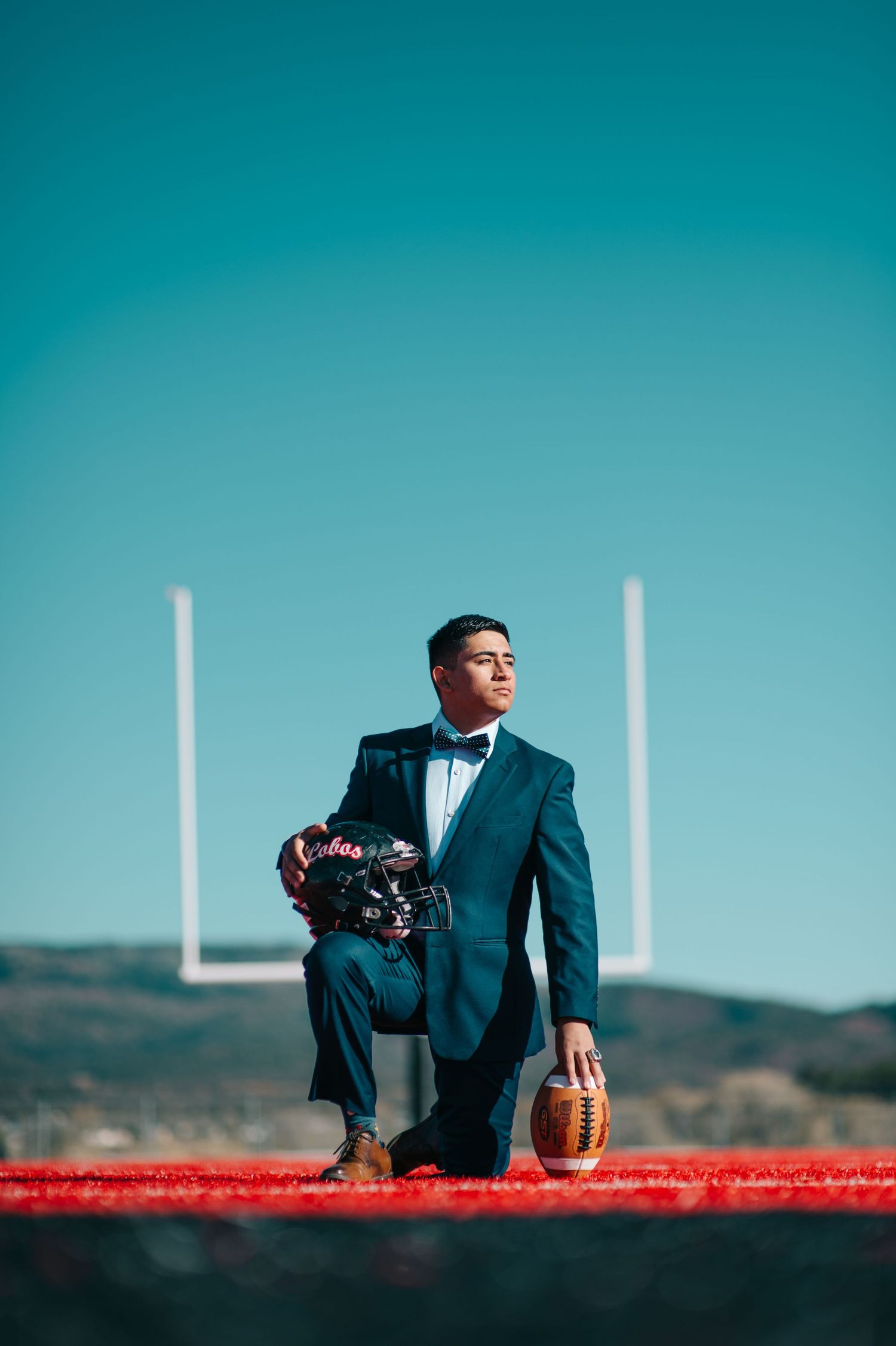 Young man in tuxedo kneeling on football field holding ball and helmet near goal post under clear blue sky.