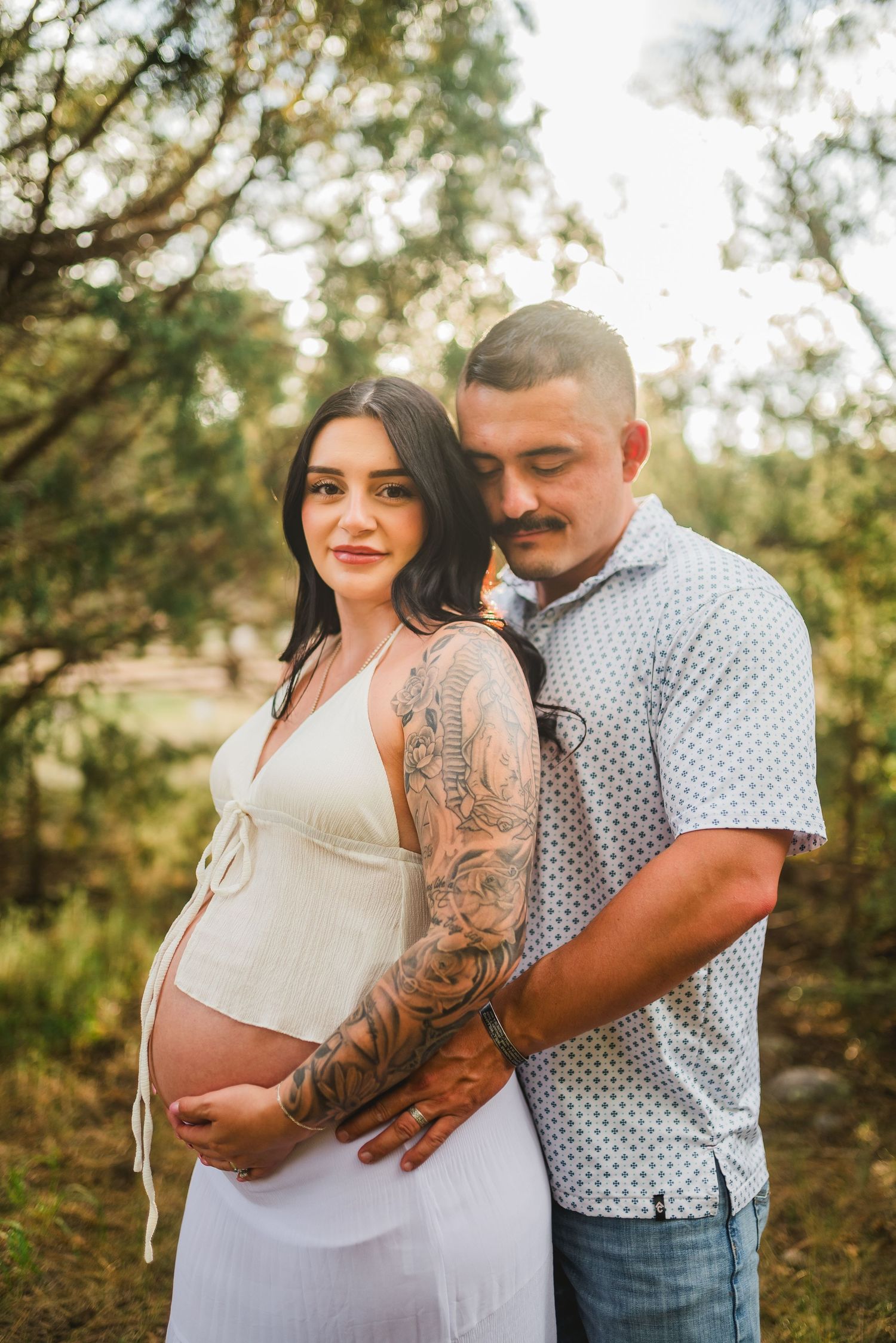 Pregnant woman in white dress with tattoos embraces partner outdoors in golden hour light during maternity photoshoot.