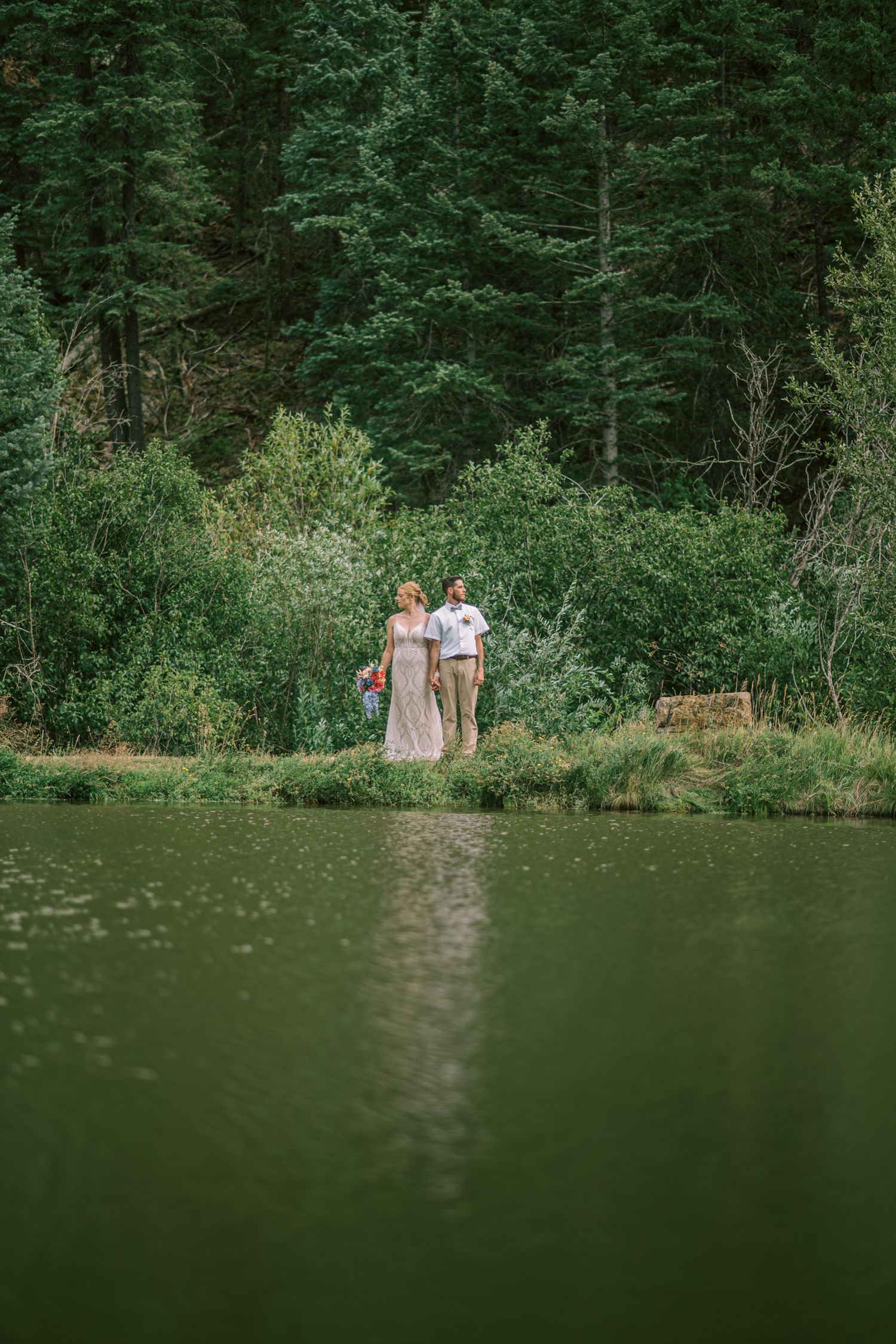 Couple standing by a serene green lake surrounded by lush evergreen forest during an outdoor maternity photoshoot.