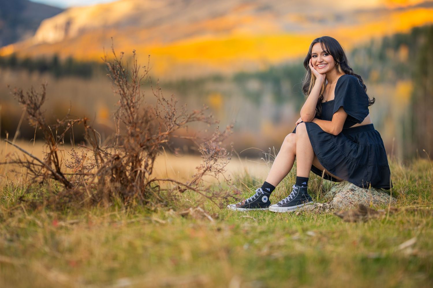 Person sits on grassy hillside during golden sunset while wearing dark clothing and looking into distance.