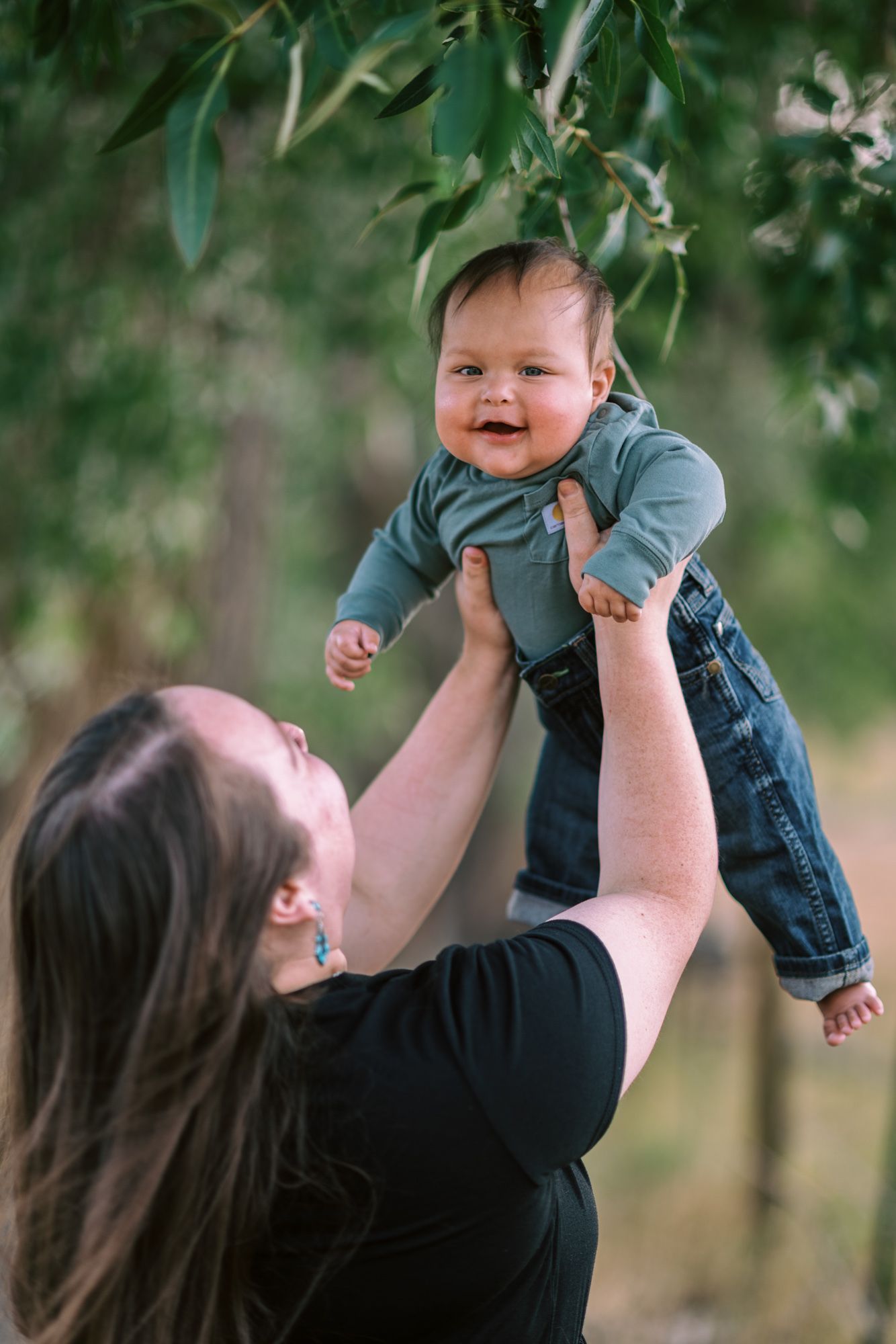 A joyful baby in denim overalls is lifted high by a mother outdoors among green trees.