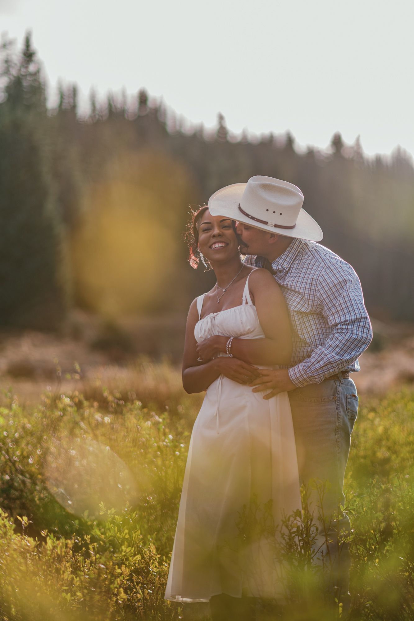 Couple embracing in sunlit meadow, man in cowboy hat kissing woman in white dress with forest backdrop.