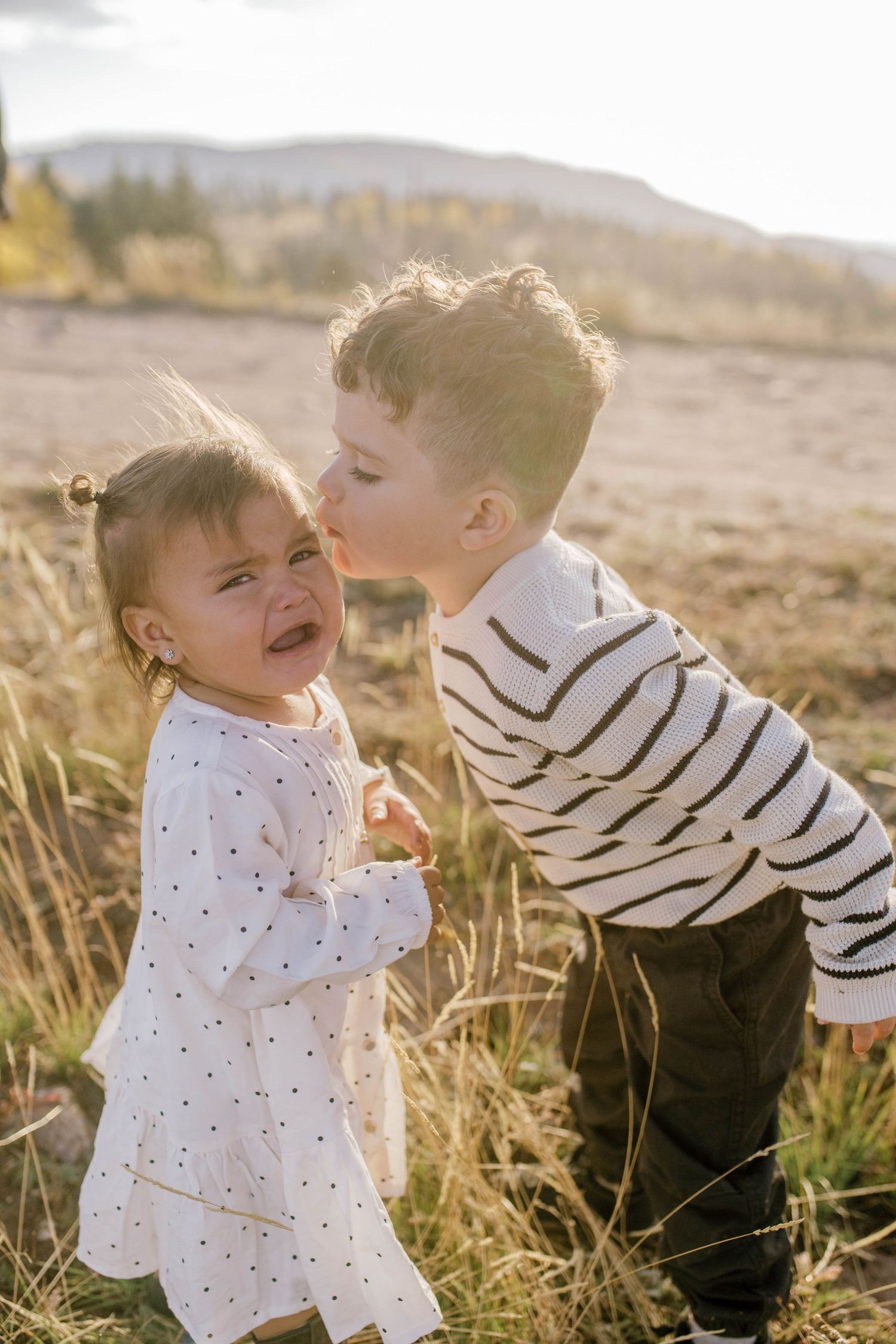 Two children share a playful moment in tall golden grass at sunset with mountains in the background.
