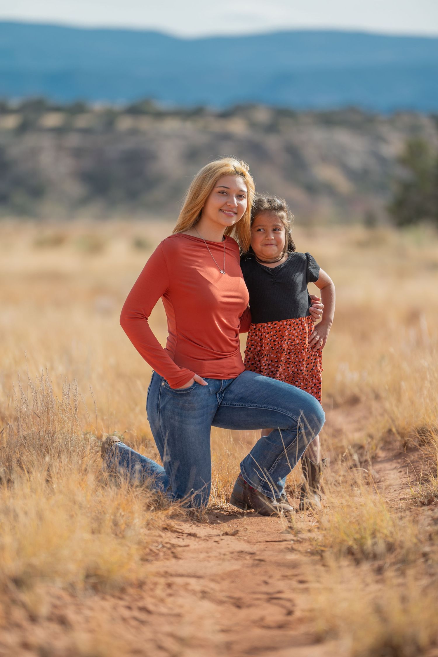 Two people pose together on a dirt trail surrounded by golden grass and mountains in the background on a sunny day.