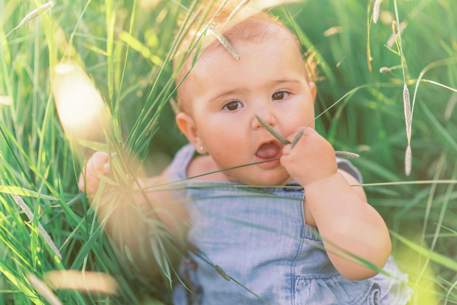 Adorable baby sitting in tall green grass, wearing denim overalls, with finger near mouth in sunny outdoor setting.