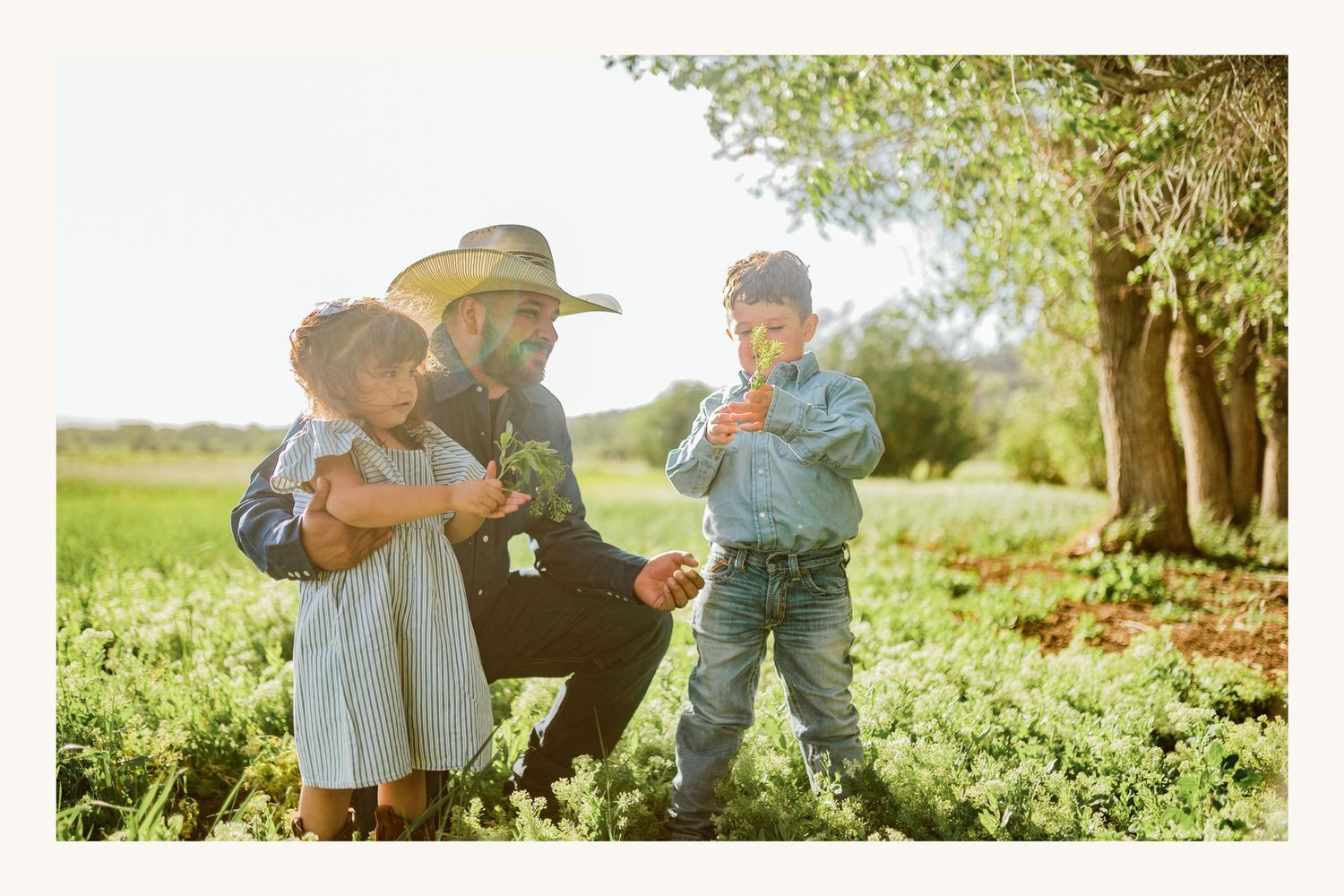 Father in straw hat crouching with two young children in a sunny green garden field.