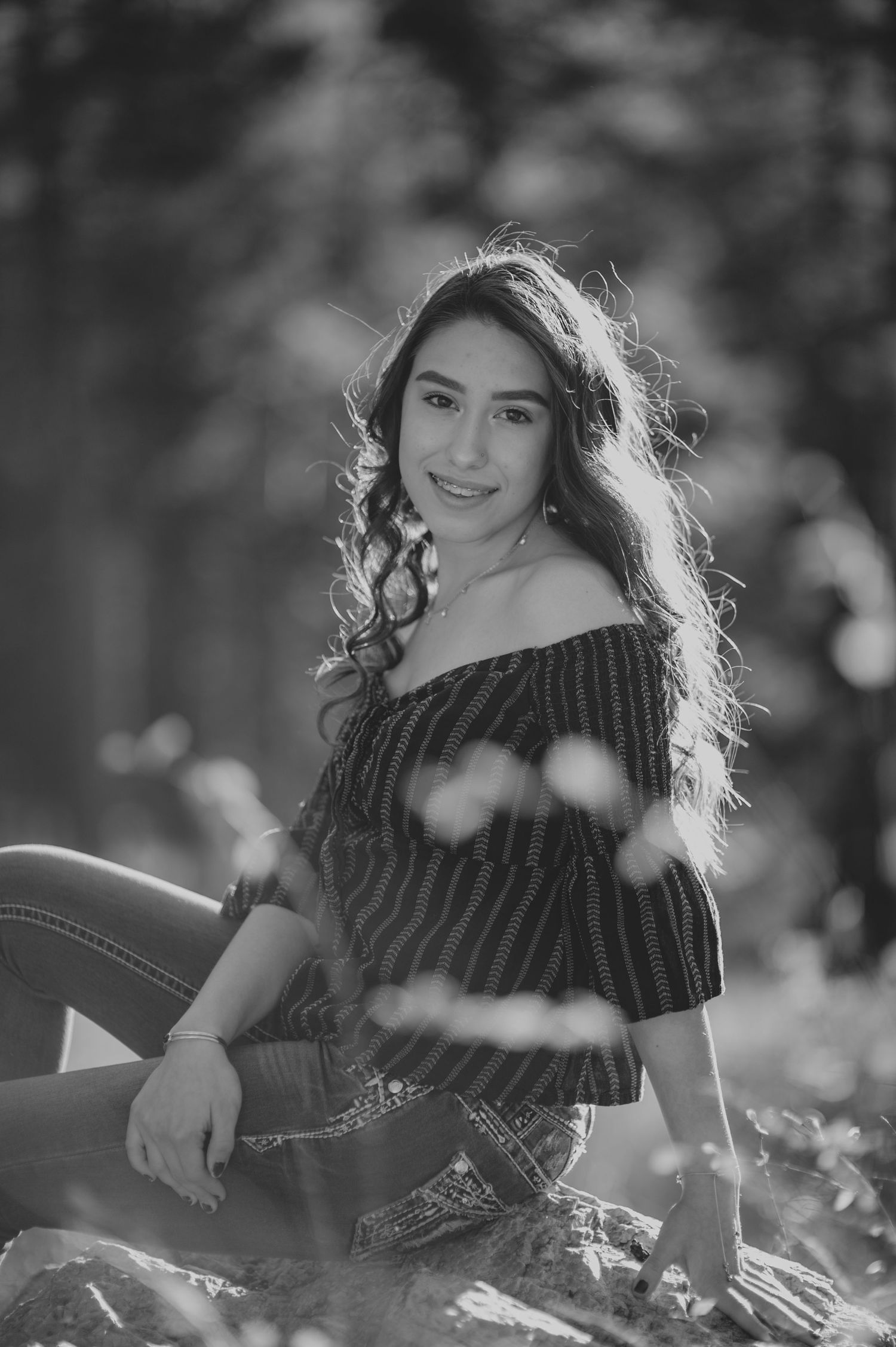 Black and white portrait of a young woman in an off-shoulder top, smiling outdoors in a sunlit forest setting.