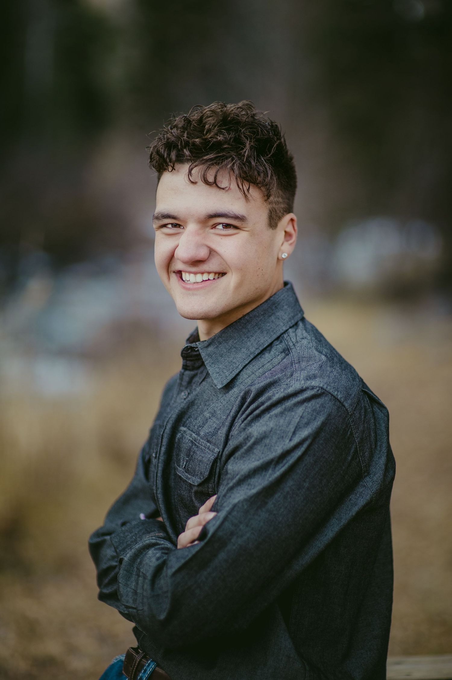Young man with curly hair smiling confidently with arms crossed, wearing a dark denim shirt outdoors.